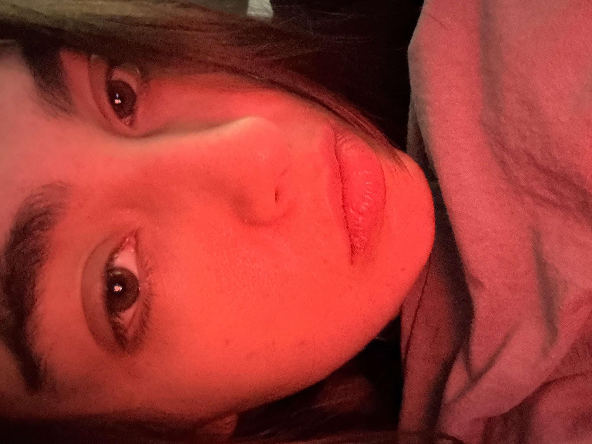 The image displays a close-up portrait of a person's face, illuminated by a dominant red light source. The subject's head is tilted significantly, appearing almost 90 degrees counter-clockwise within the frame. The person, likely a young adult, has their eyes open, with dark irises and pupils, gazing directly towards the viewer. Prominent features include thick, dark eyebrows and eyelashes, a distinct nose, and full, slightly parted lips that reflect some of the red light. Dark hair is visible, framing the upper left side of the face.
On the right side of the image, a textured, light-colored fabric, possibly part of a hoodie or blanket, is draped around the person. The background is dark and indistinct, suggesting an indoor setting in Mexico City, Mexico, likely at night or in a room with controlled lighting. There is no specific activity taking place other than the subject's direct, engaging stare. The intense red illumination creates a dramatic and intimate mood. No visible text is present in the image. The expression on the person's face appears neutral, perhaps thoughtful or observant.