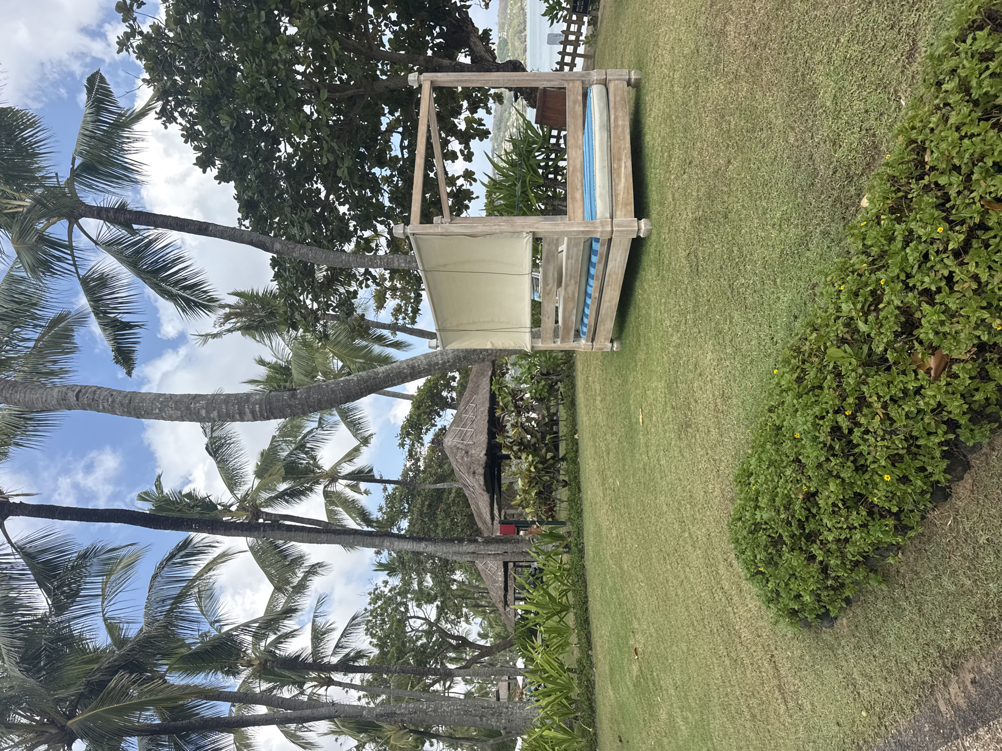The image depicts a resort scene in Kuta Selatan, Indonesia. In the foreground, a lush green lawn is dotted with small yellow flowers. A low-lying hedge runs along the bottom edge of the frame. To the right, a wooden four-poster daybed with a light-colored canopy and blue and white striped cushions sits on the grass. Behind the daybed, palm trees and other tropical vegetation are abundant. In the middle ground, two thatched-roof gazebos are visible, surrounded by greenery. Further in the background, a body of water can be seen, suggesting a coastal location. The sky is partly cloudy. There are no people visible in the frame.