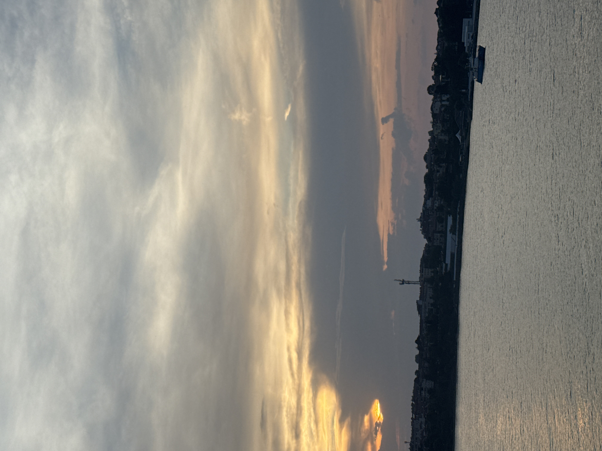 The image displays a serene coastal landscape captured during what appears to be a sunset, set in Tsarevo, Bulgaria. The scene is dominated by a vast sky and a large body of water, separated by a distant, low-lying shoreline.
The sky, occupying the upper two-thirds of the image, presents a dynamic and colorful display. Streaks of wispy, light-grey clouds blend with more substantial, darker grey formations. A prominent band of intense golden-orange and yellow light stretches horizontally across the middle of the sky, indicative of the sun's position, likely partially obscured by clouds. This warm glow contrasts sharply with the cooler, muted blues and greys of other cloud masses, creating a dramatic atmospheric effect.
Below the sky, a calm body of water, likely the Black Sea, extends into the foreground. Its surface is textured with gentle ripples, reflecting some of the sky's light, particularly from the warmer hues, as subtle streaks. The water appears predominantly dark and calm, suggesting a peaceful evening.
Along the horizon, a dark, silhouetted strip of land marks the coastline. Indistinct buildings and structures can be discerned, suggesting a coastal town or settlement. On the right side of this silhouette, a small boat or ship is visible, dark against the water, with subtle hints of lighter details on its structure. Towards the center-left of the shoreline, the distinct outline of a tall construction crane stands out, hinting at ongoing development in the area.
The overall impression is one of a tranquil twilight scene, characterized by the striking interplay of light and shadow during sunset over the sea. There are no visible people or discernible text in the image.