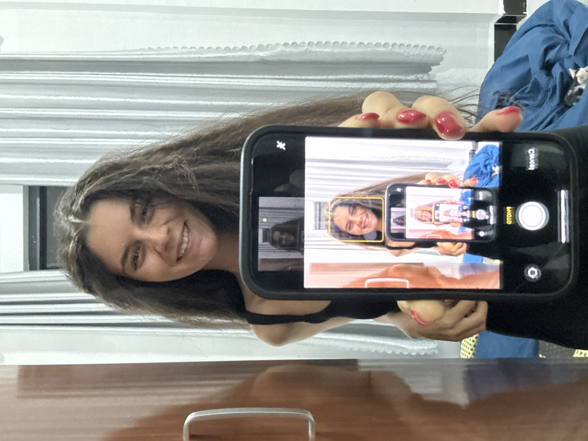 In Mũi Né, Vietnam, a young woman with long, dark, wavy hair stands holding a smartphone. She is wearing a black tank top. The phone is held out in front of her, displaying a selfie. In the selfie, the woman is smiling, and her face is framed by a yellow square, indicating face recognition. Her nails are painted a bright red. Behind her, a closet door is visible on the left, and vertical striped curtains fill the background. The scene appears to be indoors.