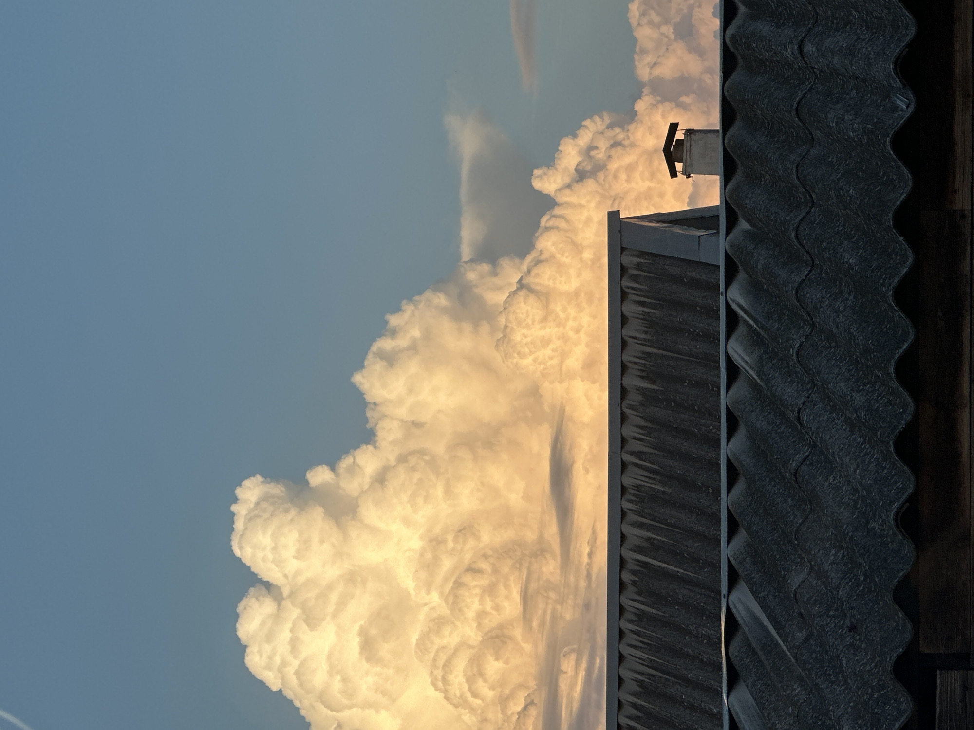 The image captures a dramatic sky scene from Primorsko-Akhtarsk, Russia, at what appears to be late afternoon or early evening, given the warm, golden light illuminating the clouds. The primary subjects are the vast, towering cumulonimbus clouds, rendered in striking shades of white and gold, hinting at their immense scale and potential for atmospheric phenomena.
A portion of a dark, corrugated roofline runs along the right side of the frame, providing a stark contrast to the ethereal clouds and grounding the scene with a sense of human presence and habitation. A small, square chimney or ventilation unit is visible atop a building in the distance, further reinforcing the impression of a residential or industrial setting. The sky itself is a clear, pale blue, creating a backdrop that emphasizes the texture and volume of the clouds.
There is no discernible activity or event taking place; the image appears to be a still moment capturing the grandeur of nature. Notable details include the powerful texture of the clouds, their illuminated edges, and the weathered appearance of the roof. The overall emotion evoked is one of awe and a sense of the sublime, juxtaposing the raw power of the weather with the mundane structures of human life. No visible text is present in the image.