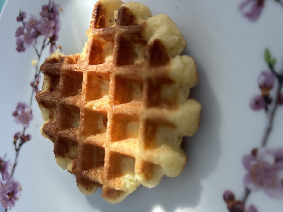 A single, golden-brown waffle with a grid pattern is centered on a white ceramic plate. A bite has been taken from the upper right edge of the waffle. The waffle displays areas of lighter yellow-brown and darker caramelization. The plate is decorated with out-of-focus pink cherry blossom-like floral branches, visible in the upper and lower background. Strong, directional lighting casts a distinct shadow from the waffle onto the plate to the lower left. No individuals, interactions, or specific environmental details beyond the plate are visible.