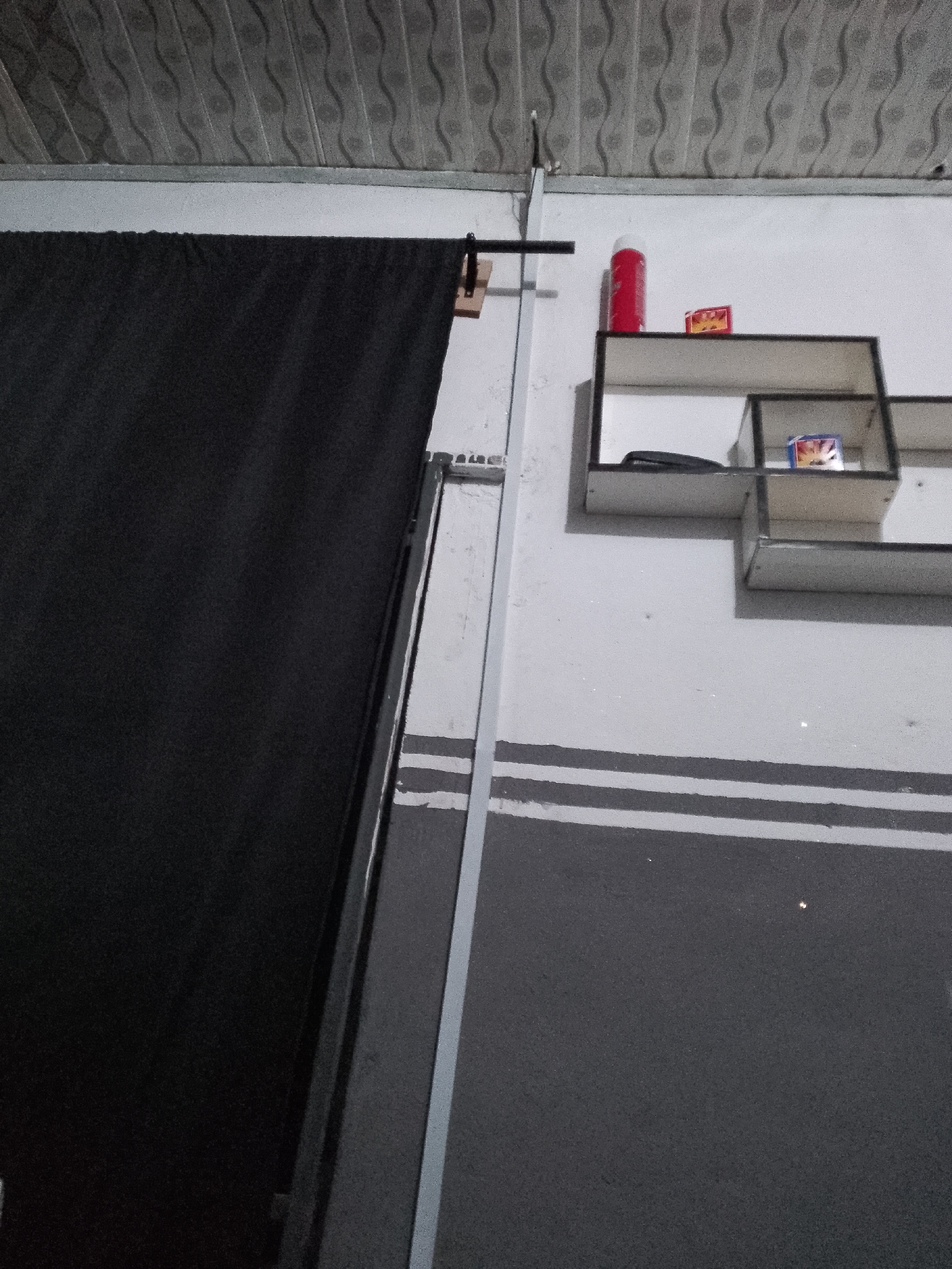 The image shows an indoor scene with a black curtain hanging from a metal rod, suspended from a ceiling that appears to be made of corrugated panels. To the right of the curtain, a white wall is visible, featuring a set of three wall-mounted shelves. On the top shelf, a red cylindrical object, possibly a can, and a small rectangular box are placed. The middle shelf holds a dark object, possibly a wallet or phone, and another small rectangular box with a colorful design. The bottom shelf is empty. Below the shelves, the wall is painted with two horizontal dark gray stripes. A vertical metal pole runs along the wall, adjacent to the curtain. The floor is not visible. The overall impression is of a sparsely decorated room, potentially a storage area or a makeshift studio. The location in Jauro Yinu, Nigeria, suggests a context of a local, possibly residential or small commercial, environment.