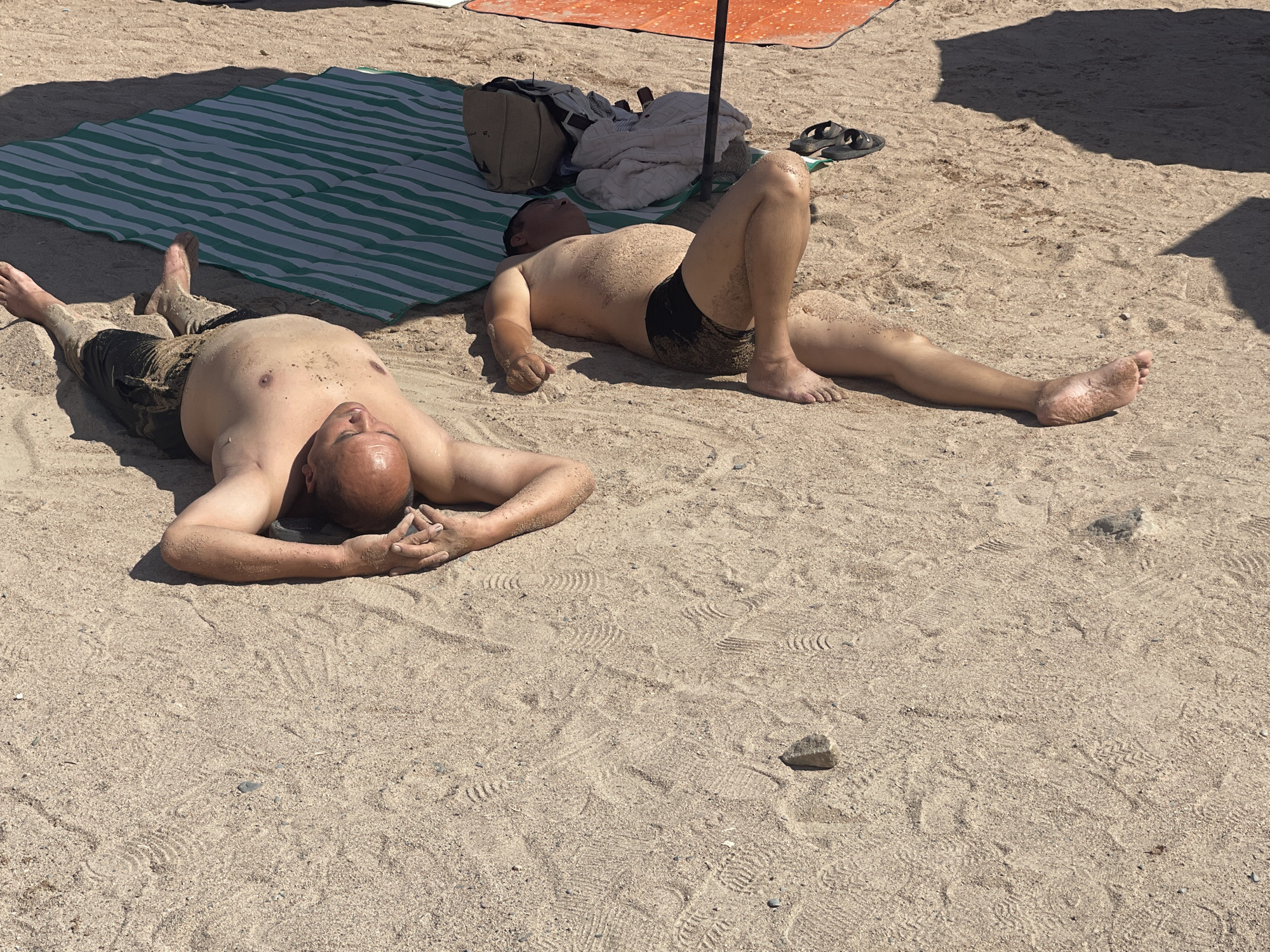 Two men are lying on a sandy beach, sunbathing. The man in the foreground on the left is lying on his back with his arms behind his head, wearing dark shorts. The man on the right is lying on his side with his legs extended, wearing black swim trunks. A green and white striped mat is laid out behind the man on the left, and an orange umbrella is partially visible in the background. The beach appears to be located in Bosteri, Kyrgyzstan.