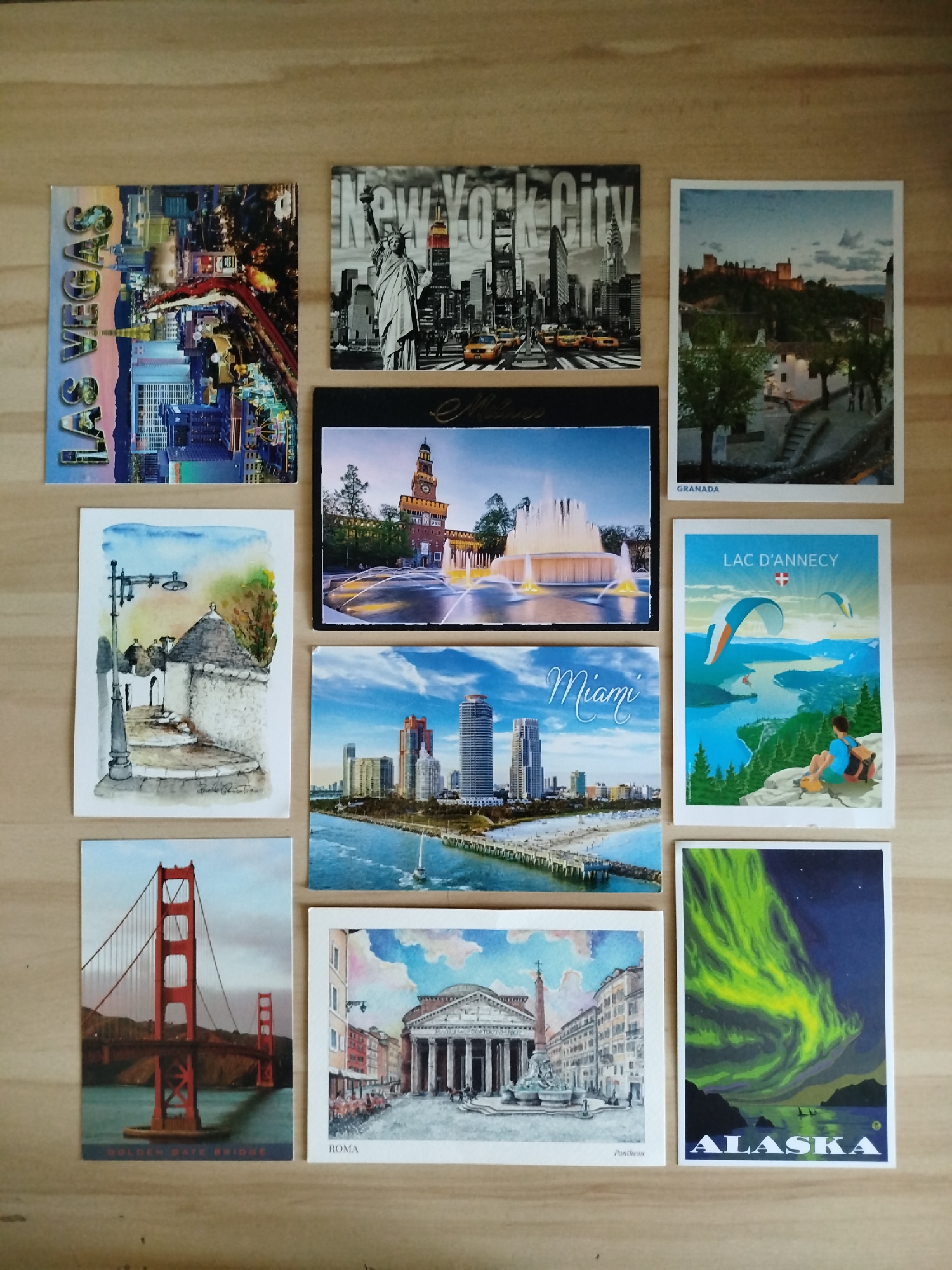 The image shows a collection of postcards arranged on a wooden surface. The postcards feature various locations around the world. Clockwise from top left: Las Vegas, New York City, Granada, Lac D'Annecy, Alaska, Roma (Pantheon), Golden Gate Bridge, Miami, Alberobello (Italy), Milan. There is no scene depicting Varna, Bulgaria.