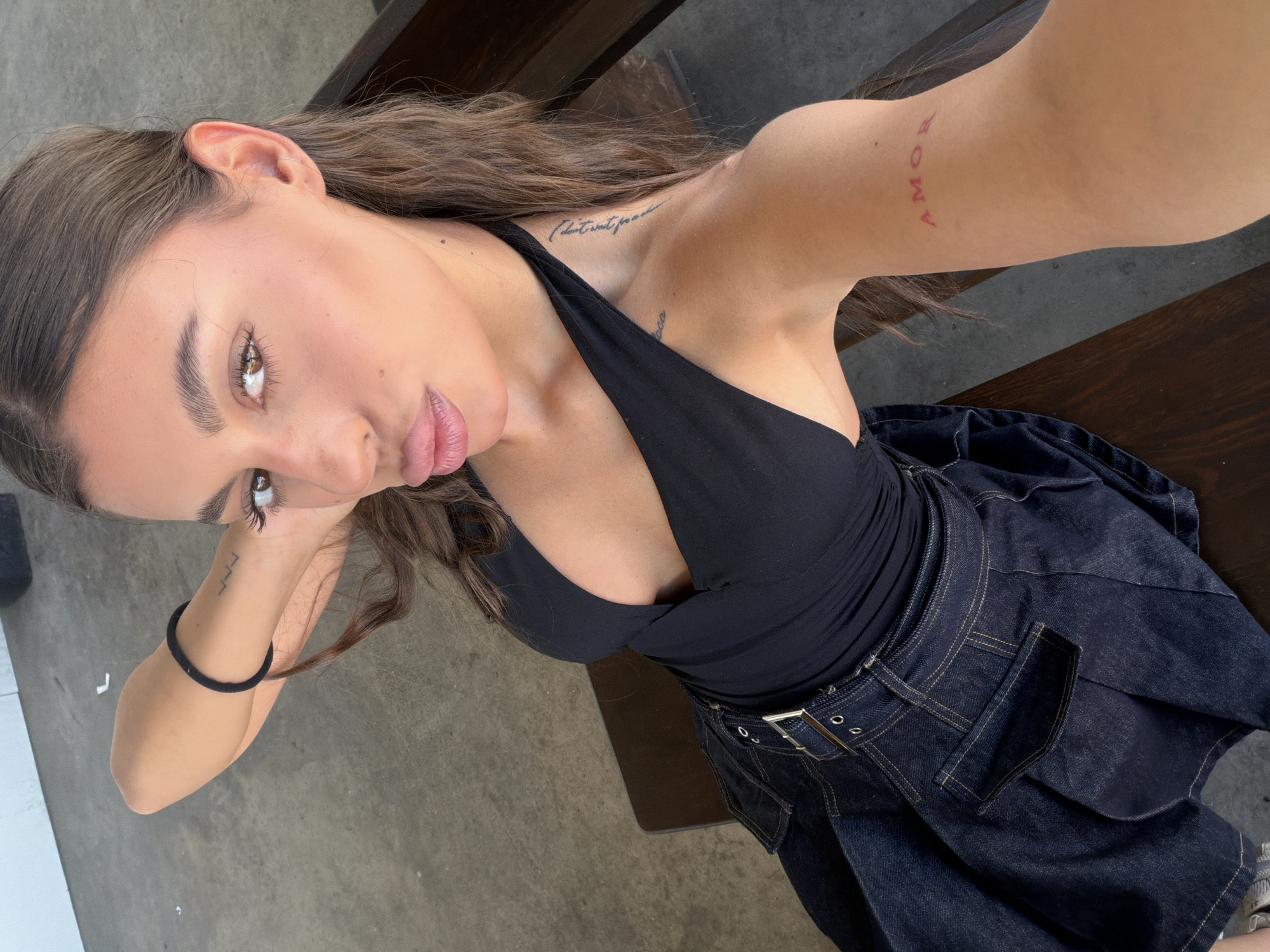 A young woman with long brown hair, wearing a black halter top and a dark denim skirt, poses for a selfie. She is seated on a wooden bench outdoors, with a concrete floor visible. The woman has several tattoos, including the word "AMOR" in red on her upper arm. The image has been taken in Samovodene, Bulgaria.