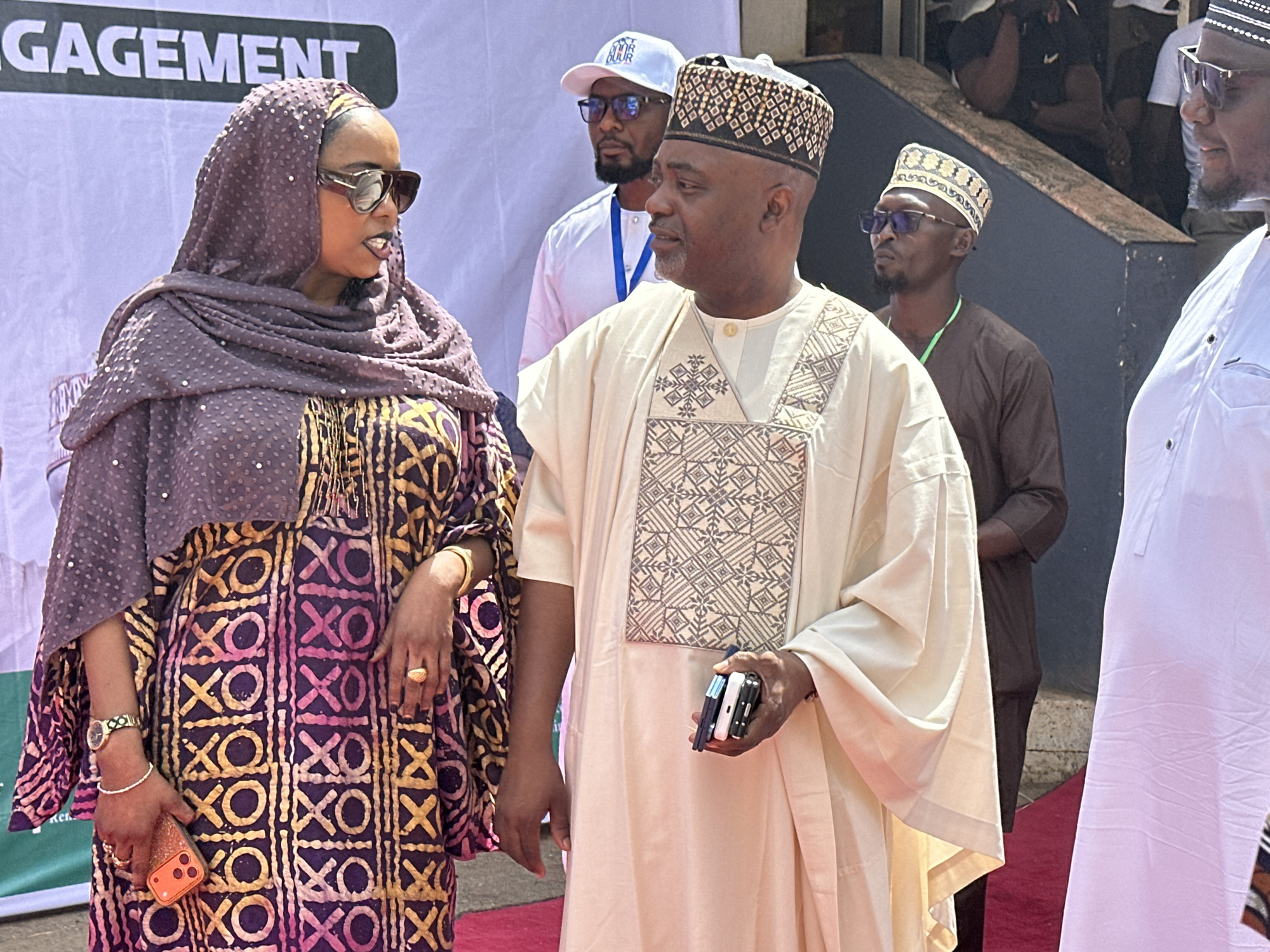 The image shows a group of people, two of whom are prominent in the foreground. A woman, wearing a purple headscarf and large sunglasses, stands next to a man wearing a traditional patterned hat and robe. The woman holds a phone in her left hand. The man holds three phones in his left hand. There are three other men in the background. The setting seems to be a formal event, as indicated by the backdrop with "ENGAGEMENT" visible. The exact location within the city of Unknown, Unknown cannot be determined from this image.