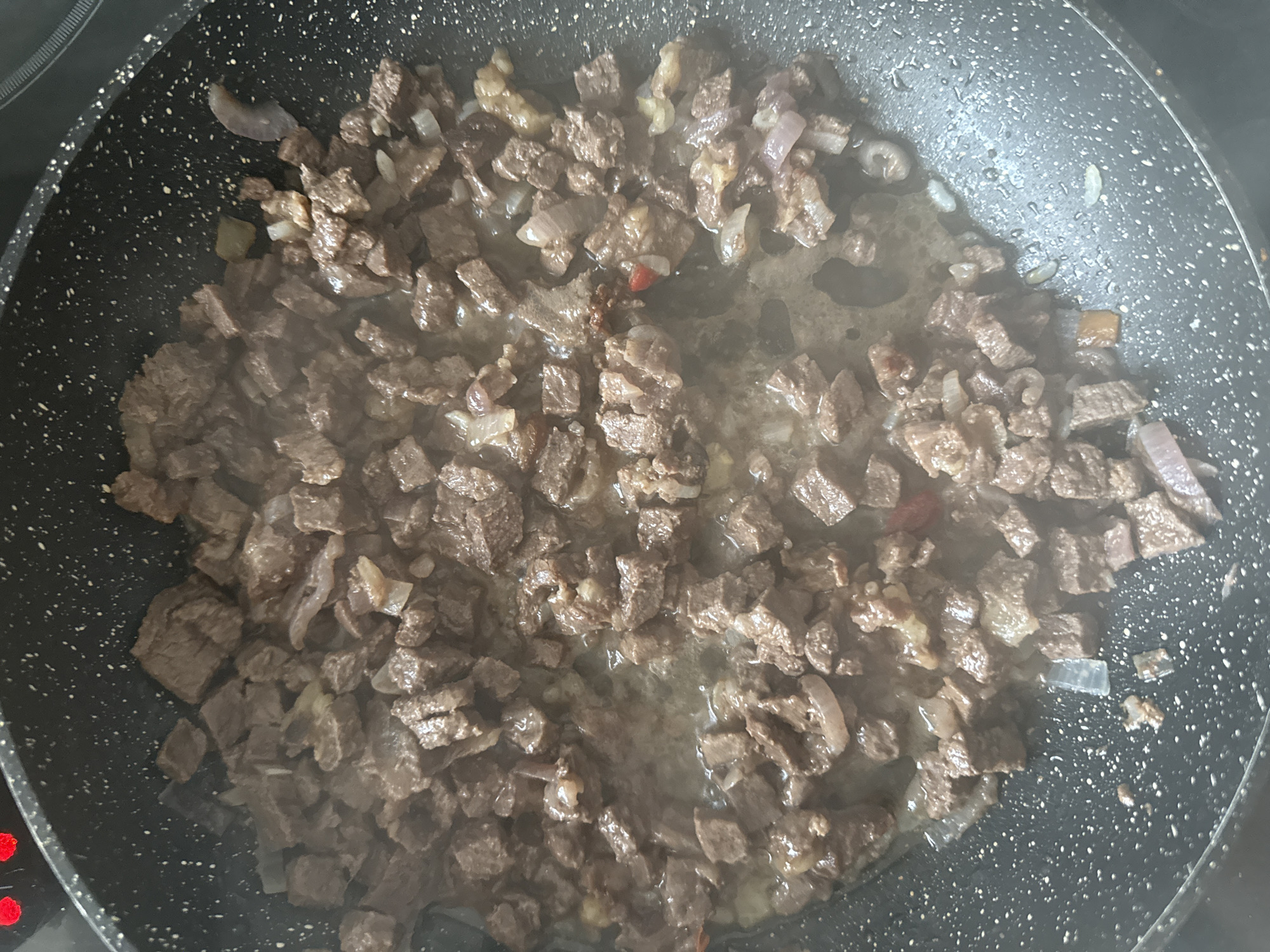 The image shows a close-up, overhead view of diced beef and onions being cooked in a black, speckled frying pan. Steam is rising from the pan, indicating that the food is hot and actively cooking. The beef is browned and appears to be in small, irregular cubes. The onions are also diced and interspersed with the meat, some pieces appearing translucent from cooking. There is a small amount of liquid, likely rendered fat or juices, at the bottom of the pan. The background is dark, suggesting the pan is on a stovetop. There is no visible evidence to suggest this image is located in Yakutsk, Russia; the scene depicts a common cooking scenario.