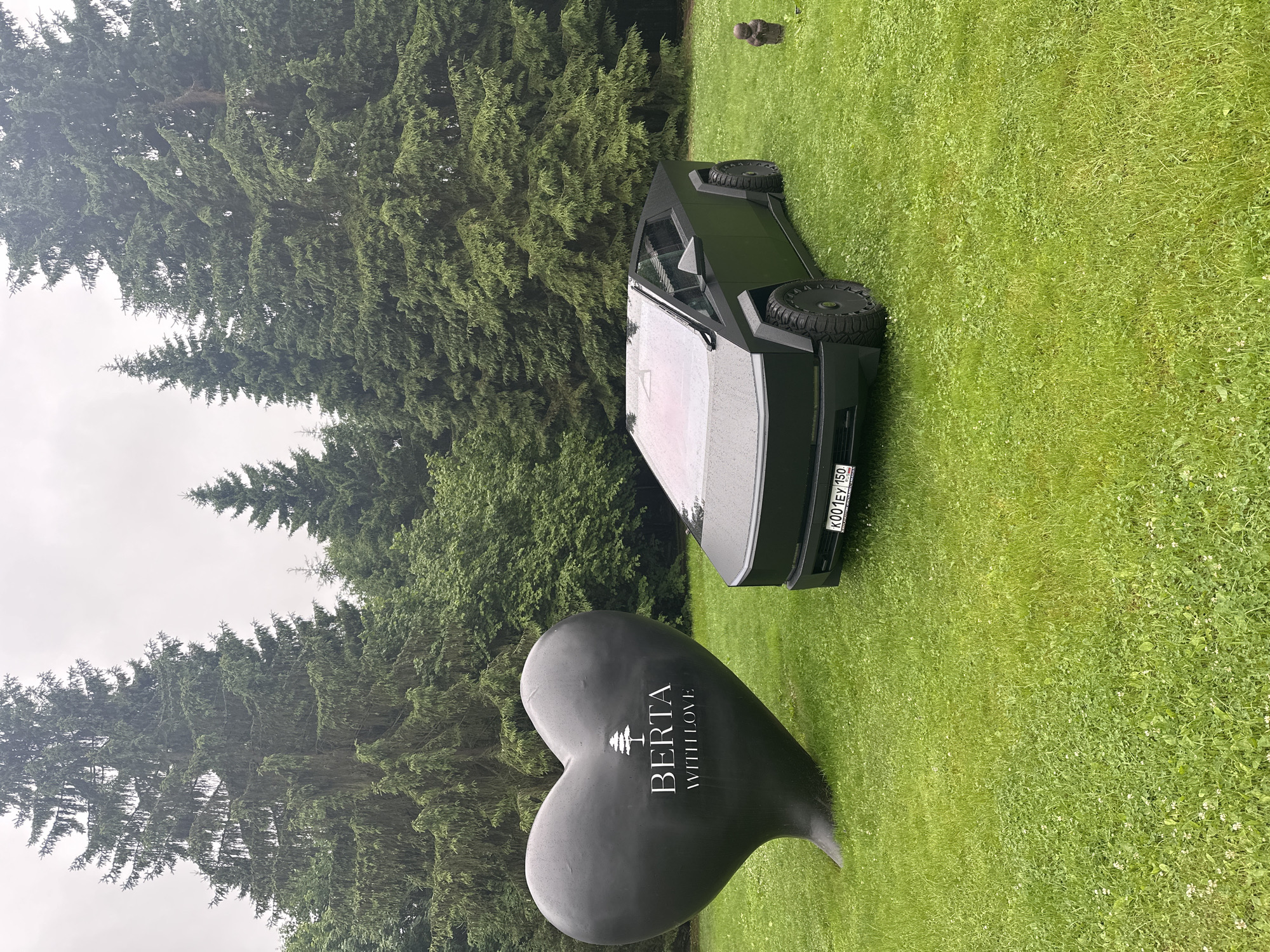 An outdoor scene features a black Tesla Cybertruck parked on a green, grassy lawn. To the left of the vehicle is a large, black, heart-shaped sculpture with white lettering that reads "BERTA WITH LOVE" and a small tree emblem. In the background, a dense forest of tall evergreen trees with a slightly overcast sky can be seen. A small, dark statue of a figure is visible on the right side of the frame. The license plate on the Cybertruck is visible and reads "K001EY 150". The location is посёлок гидроузла имени Куйбышева, Russia.