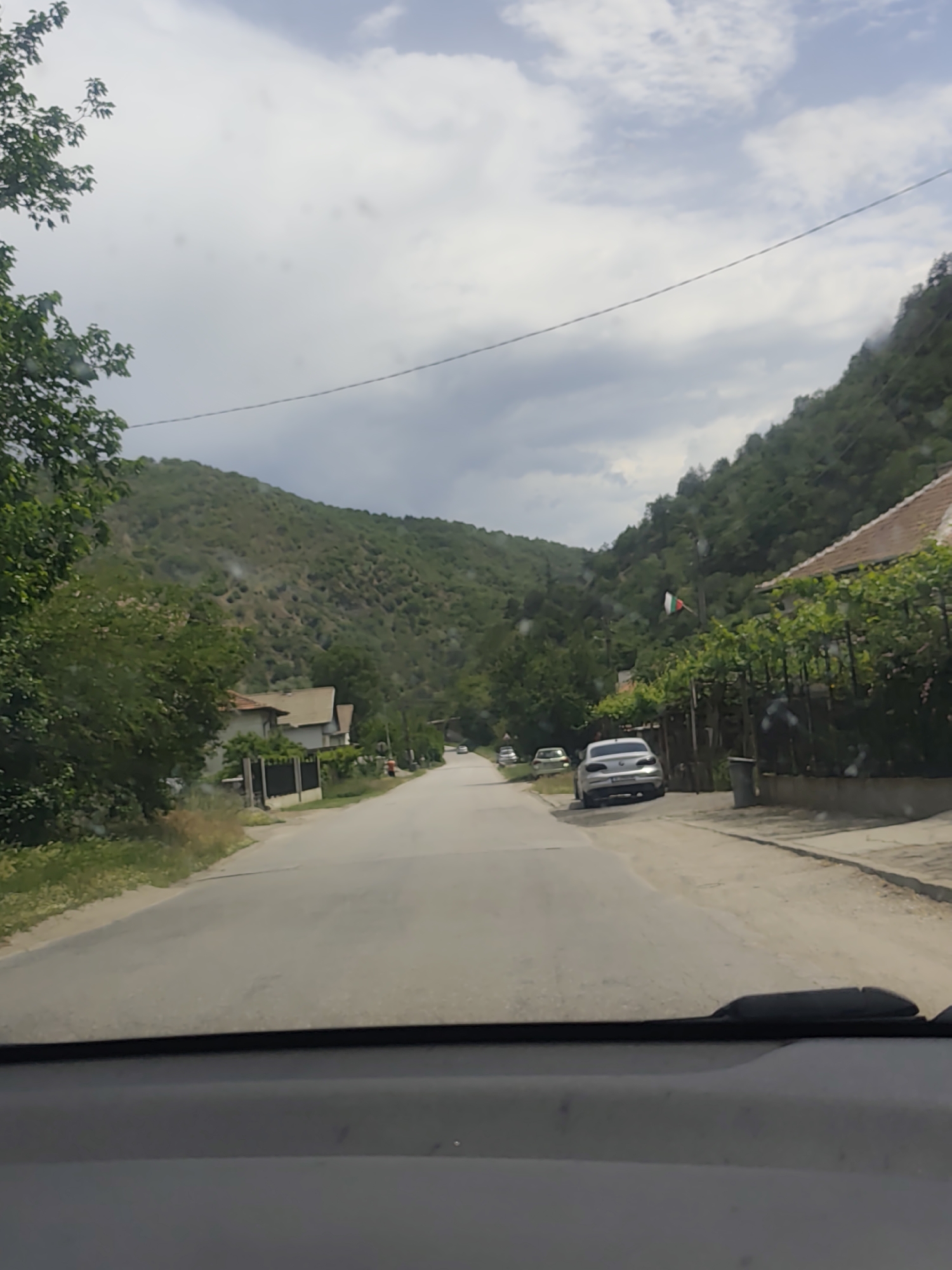 The image depicts a street scene in Jeleznitsa, Bulgaria, as viewed from inside a car. The road stretches into the distance, flanked by houses and lush greenery. Mountains rise on either side of the road. A silver car is parked on the right side of the road. A Bulgarian flag flutters near one of the houses. The sky is overcast with cloudy. The car's dashboard and windshield wiper are visible at the bottom of the frame.
