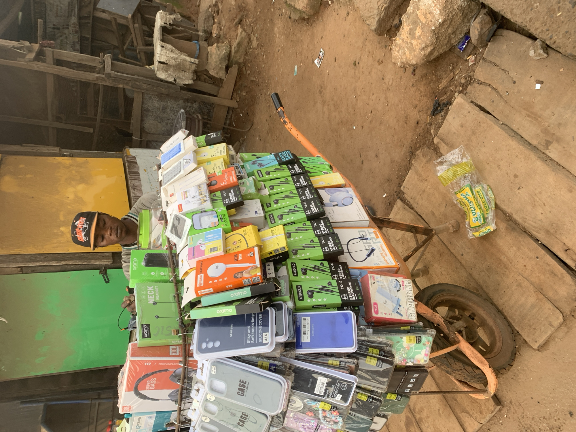 In Shagamu, Nigeria, a young man wearing a baseball cap with an orange logo stands behind a cart filled with electronic accessories. The cart, painted orange, is laden with phone cases, headphones, chargers, and other mobile accessories displayed in colorful packaging. Behind the cart, a makeshift stall constructed from wood and corrugated metal panels painted green and yellow serves as a backdrop. The ground is covered with dirt and wooden planks, with scattered debris and rocks. A person is operating a mobile cart laden with accessories for sale in a street market environment.