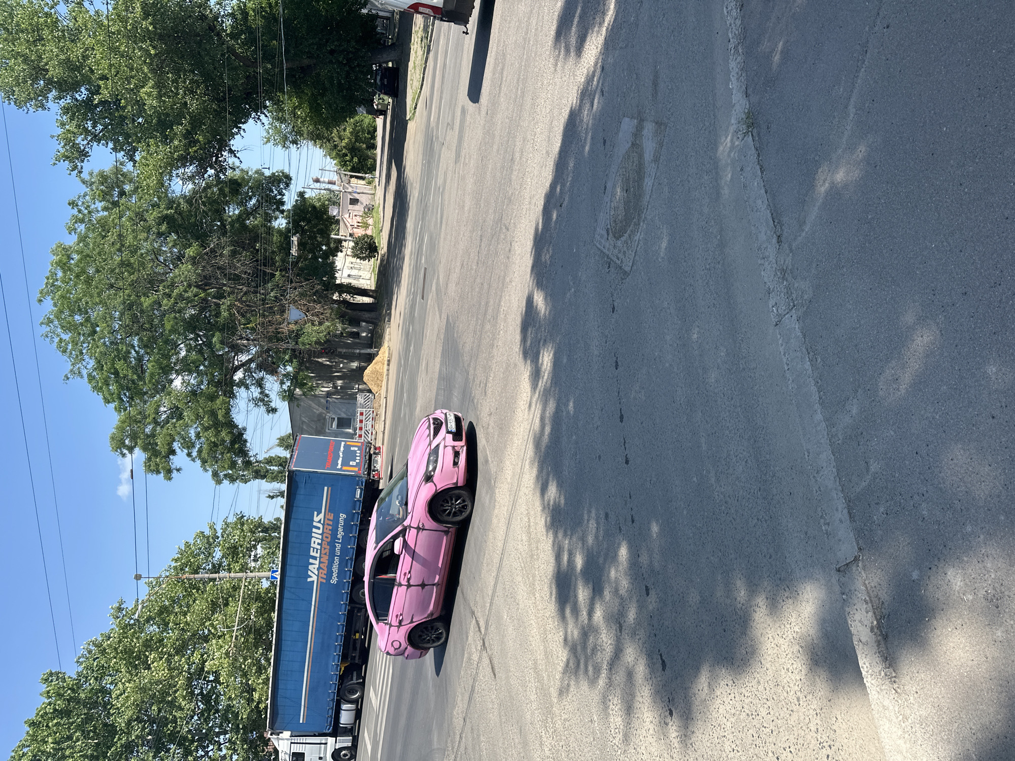 A pink sedan is driving through an intersection on a sunny day in an urban environment. A large blue semi-truck with "VALERIUS TRANSPORTE" written on its side is stopped at the intersection. The road is asphalt, and there are trees and buildings in the background. The sky is clear and blue.