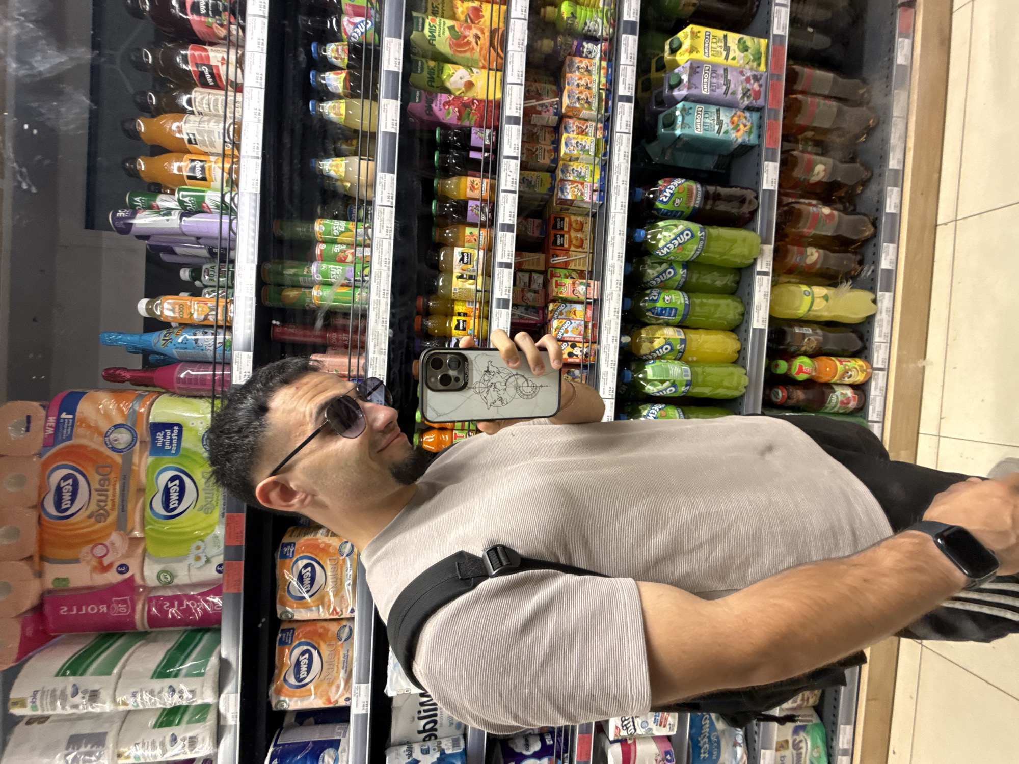 A man is taking a selfie in a supermarket aisle. He is wearing a grey t-shirt, black shorts, and a black backpack. He is also wearing sunglasses and a smartwatch. The shelves behind him are stocked with various food and drink items, including toilet paper, juices, and bottled drinks. The location is a supermarket in Varna, Bulgaria. The man is looking at his phone, which is held in his right hand. The phone has a case with a world map design. The lighting in the supermarket is bright and artificial. The floor is tiled.