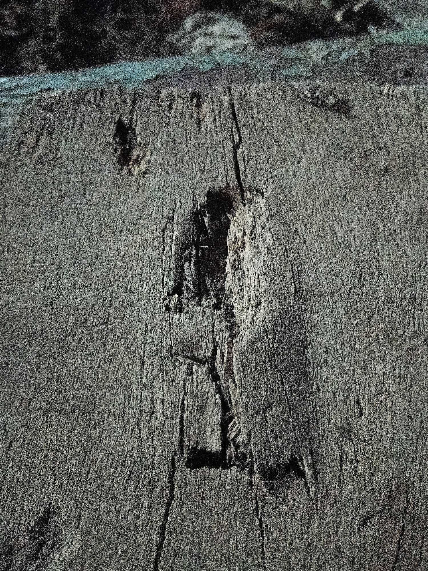 The image shows a close-up, low-angle shot of a weathered, gray wooden surface. The wood exhibits a rough texture with visible grain lines and several imperfections, including a large, irregular hole or cavity in the center. This cavity appears to be a gouge or a section of rot, revealing darker, rougher material within. There are also vertical cracks running through the wood. The top edge of the wooden surface is visible, with a lighter, bluish-gray band above it, possibly representing a transition to another material or a different lighting condition. The background above this band is dark and out of focus, with hints of brown and green. The lighting creates subtle shadows that highlight the texture and unevenness of the wood. There are no people, animals, or any discernible signs of human activity or interaction in this image. The image does not provide any information that would allow for the determination of its location in Cikal, Indonesia, or any other specific geographical location.