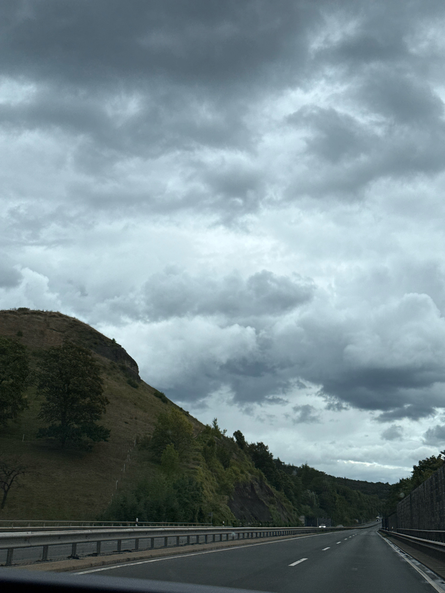 The image depicts a highway with a guardrail on the left and a wooded hillside on the right. The sky is heavily overcast with dark, dramatic clouds. The road appears to be in the vicinity of Herzberg, Germany, given the context provided. There are no people visible in the image. The scene is captured from the perspective of someone traveling by car on the highway.