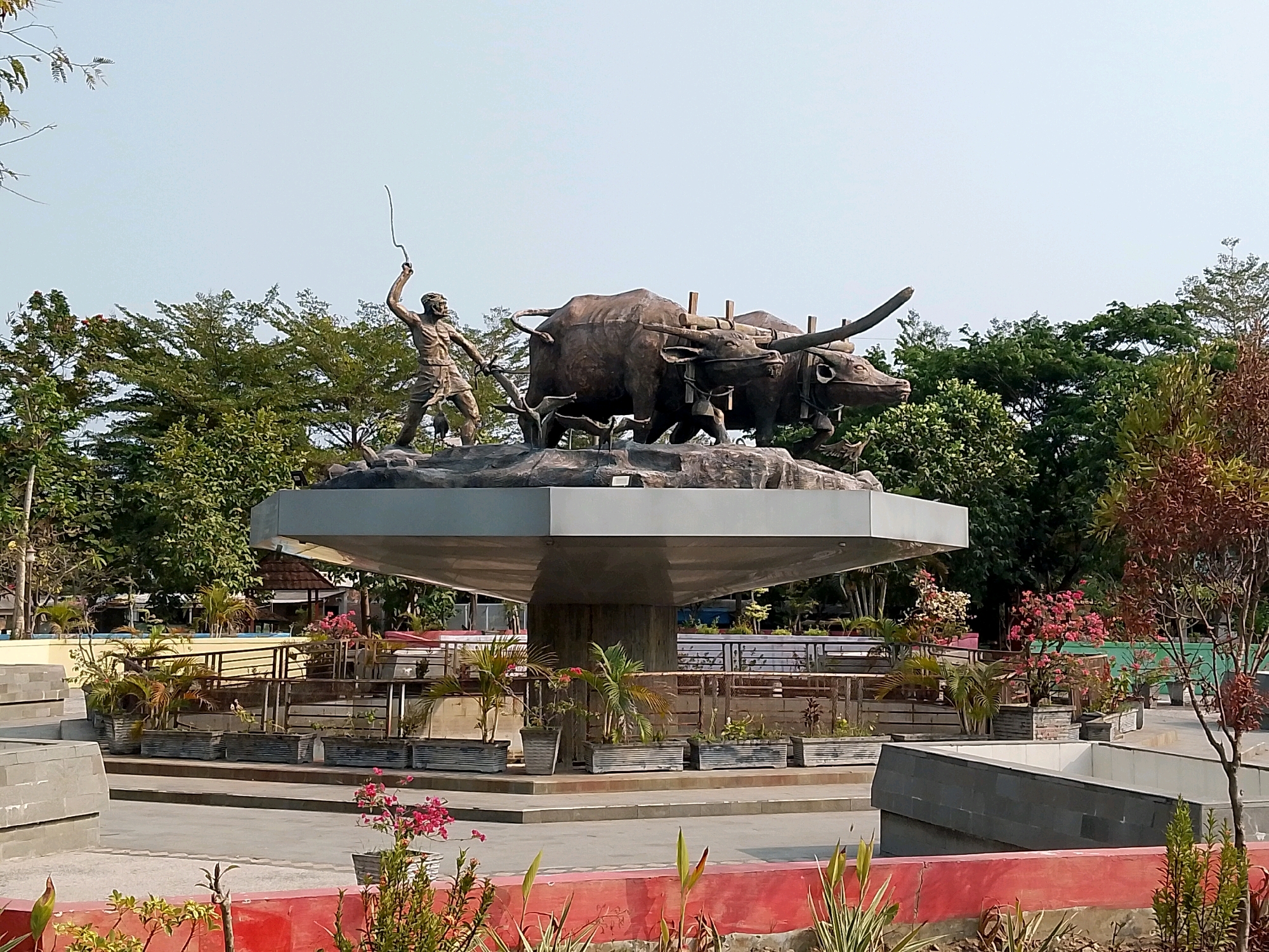 The image depicts a bronze statue of a farmer with two water buffaloes pulling a plow. The statue is situated on a circular concrete platform in a public space, likely a park or monument. The farmer, depicted in a dynamic pose, holds a whip in his raised right hand, guiding the water buffaloes forward. The buffaloes are depicted with their yokes and plow.
The statue is surrounded by lush greenery, including various trees and potted plants. In the background, there are glimpses of buildings and fences, suggesting an urban or semi-urban setting. The overall ambiance is that of a monument celebrating agricultural heritage.
This statue is a prominent landmark in the city of Ngawi, Indonesia, and is known as the "Sasono Plaza" or "Plaza Sasono." It symbolizes the agricultural prowess of the region and serves as a gathering point for locals and tourists alike. The statue's realistic depiction of traditional farming practices evokes a sense of history and cultural pride. The surrounding area is well-maintained, with paved walkways and seating areas, making it an inviting space for recreation and reflection.