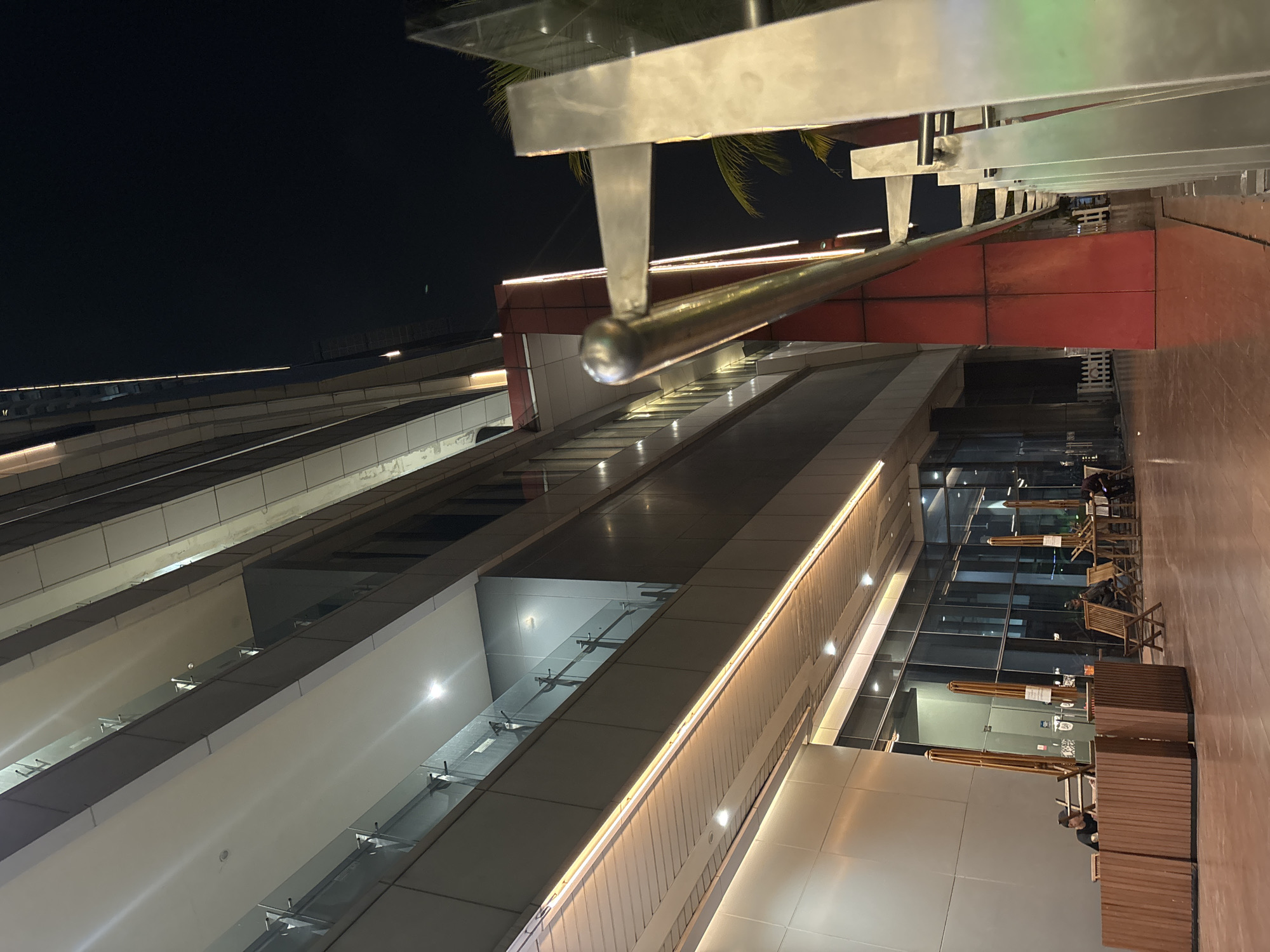 The image shows a nighttime view of a modern building facade in North Jakarta, Indonesia. The building features multiple levels with glass railings on balconies and vertical strip lighting accentuating the architectural design. In the foreground, a polished metal railing runs along a walkway. To the left, there are outdoor seating areas with wooden tables and chairs, some with umbrellas, and a few individuals are seated or present in the area. The ground appears to be a reddish-brown tiled or paved surface. The sky is dark and clear.
