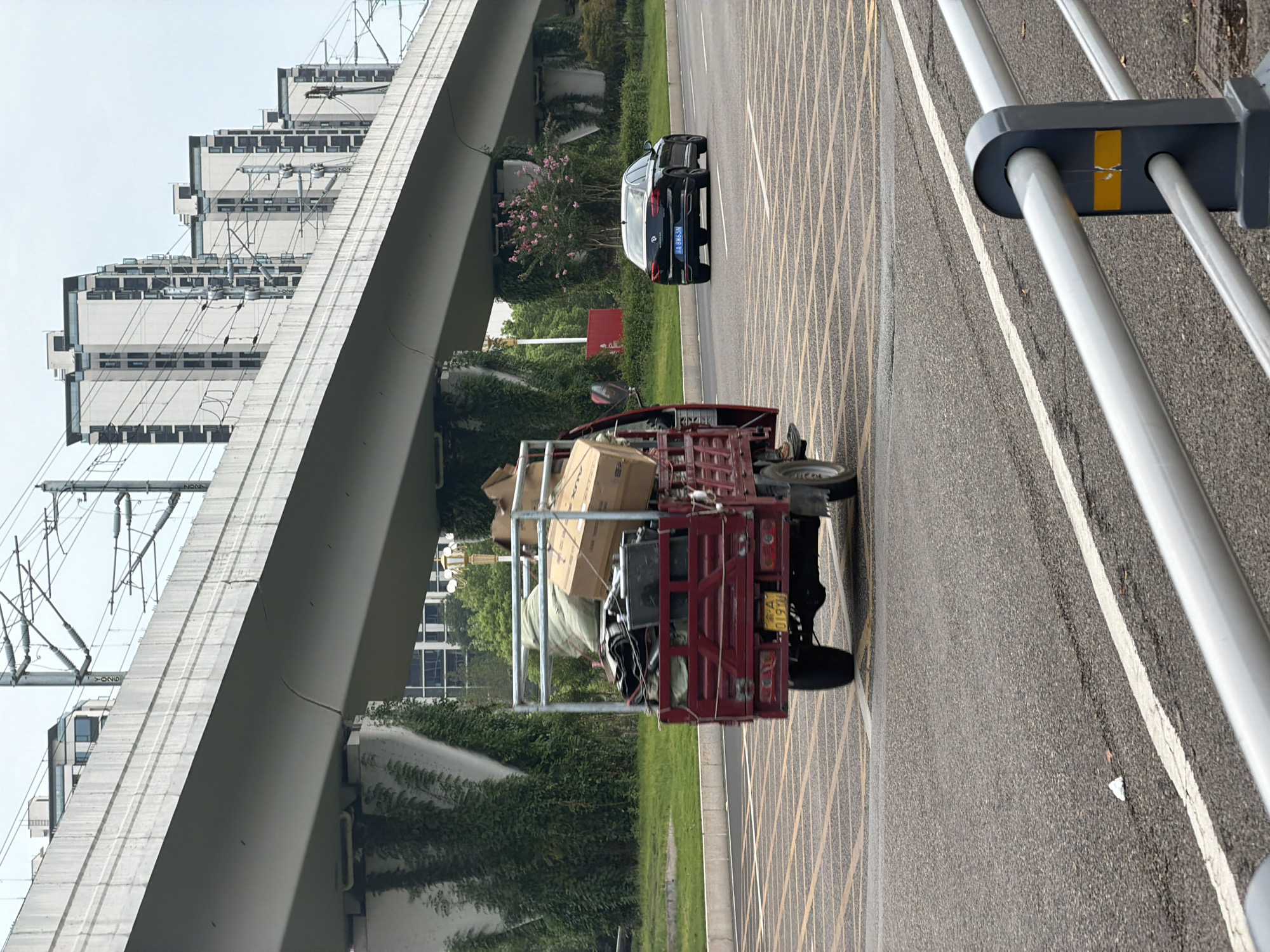 A red flatbed truck is driving on a road in the city of Jiangjin, China. The truck is loaded with boxes and bags. A black sedan is driving on the road behind the truck. A large elevated highway structure is overhead. Buildings and trees are visible in the background. The sky is overcast.
