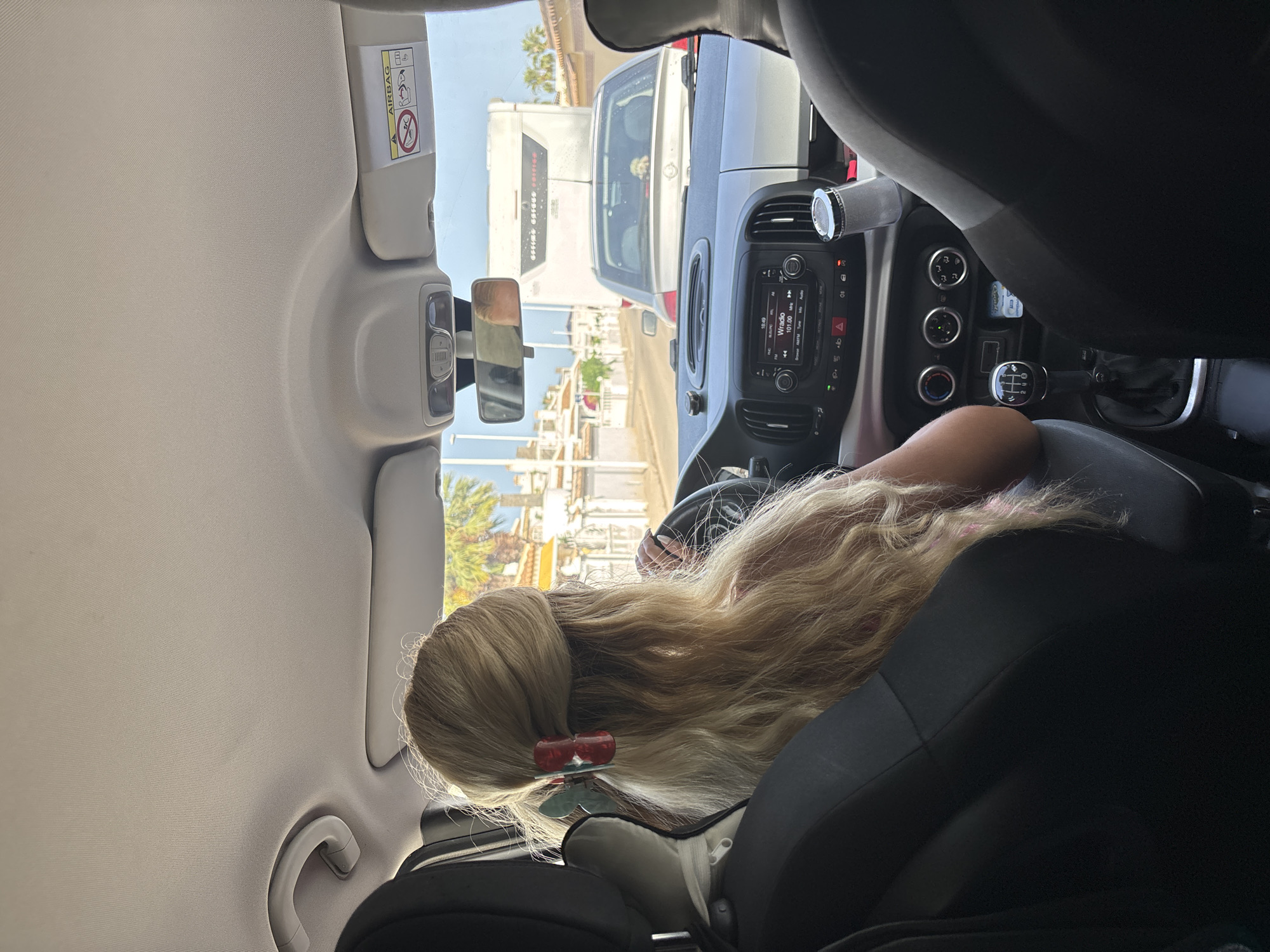 This image, taken inside a car in Orihuela, Spain, shows a woman with blonde hair, styled in a half-updo with a decorative clip, driving. She is wearing a pink top and has her hands on the steering wheel. The car's interior is visible, including the dashboard with a radio display showing "Wradio 101.00 MHz", air vents, and a manual gear stick. Outside the car, a white camper van and a silver car are ahead in traffic on a street lined with houses and palm trees, typical of the Orihuela region. The sky is clear and blue, indicating a sunny day.
