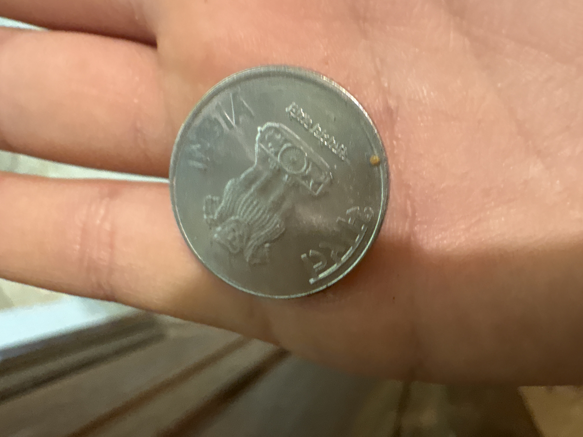 A close-up, overhead shot shows a person's palm holding a silver-colored coin. The coin is positioned in the center of the palm and is slightly tilted. The coin appears to be an Indian rupee coin, with a depiction of the Ashoka pillar and Indian script visible. The palm is lightly tanned and shows the natural lines and texture of skin. In the upper right quadrant of the coin, the word "INDIA" is partially visible in raised lettering. Below the Ashoka pillar, more Indian script is present. There is a tiny, light brown speck on the lower left edge of the coin. The background consists of blurred, vertical wooden or painted surfaces, suggesting an indoor setting. The lighting is soft, casting a gentle illumination on the coin and palm. The scene is static, with no visible action or interaction occurring. The location is not specified in the image.