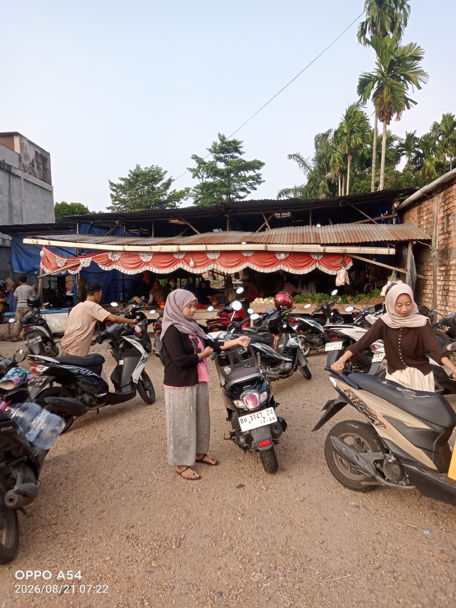 The image captures a street scene in Jambi, Indonesia, possibly near a local market. Two women, one in a light pink hijab and patterned pants, and another in a brown hijab and pants, stand near motorcycles. The woman in pink is adjusting her stance, while the woman in brown is looking down. A man in a beige t-shirt and pants is interacting with a white and black motorcycle, appearing to be preparing to ride it. The background features a covered stall with red and white fabric draping, suggesting it's a vendor area. Various motorcycles are parked around the scene, and the ground is covered in gravel. Palm trees and other greenery are visible in the background, along with a brick building and parts of a concrete structure. The lighting suggests it is either early morning or late afternoon.