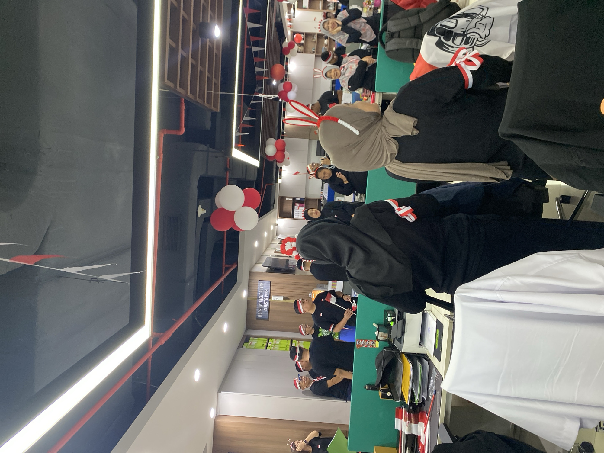 The image depicts an office interior decorated with red and white Indonesian flags and balloons. Several individuals, dressed in black attire and adorned with red and white headbands and armbands, are gathered in the space. Some are holding small Indonesian flags. The environment appears to be a workspace with cubicles, desks, chairs, and office equipment visible. The ceiling has prominent lighting fixtures and exposed conduits. The overall scene suggests a celebration or event within an office setting in Central Jakarta, Indonesia, likely related to Indonesian national holidays.