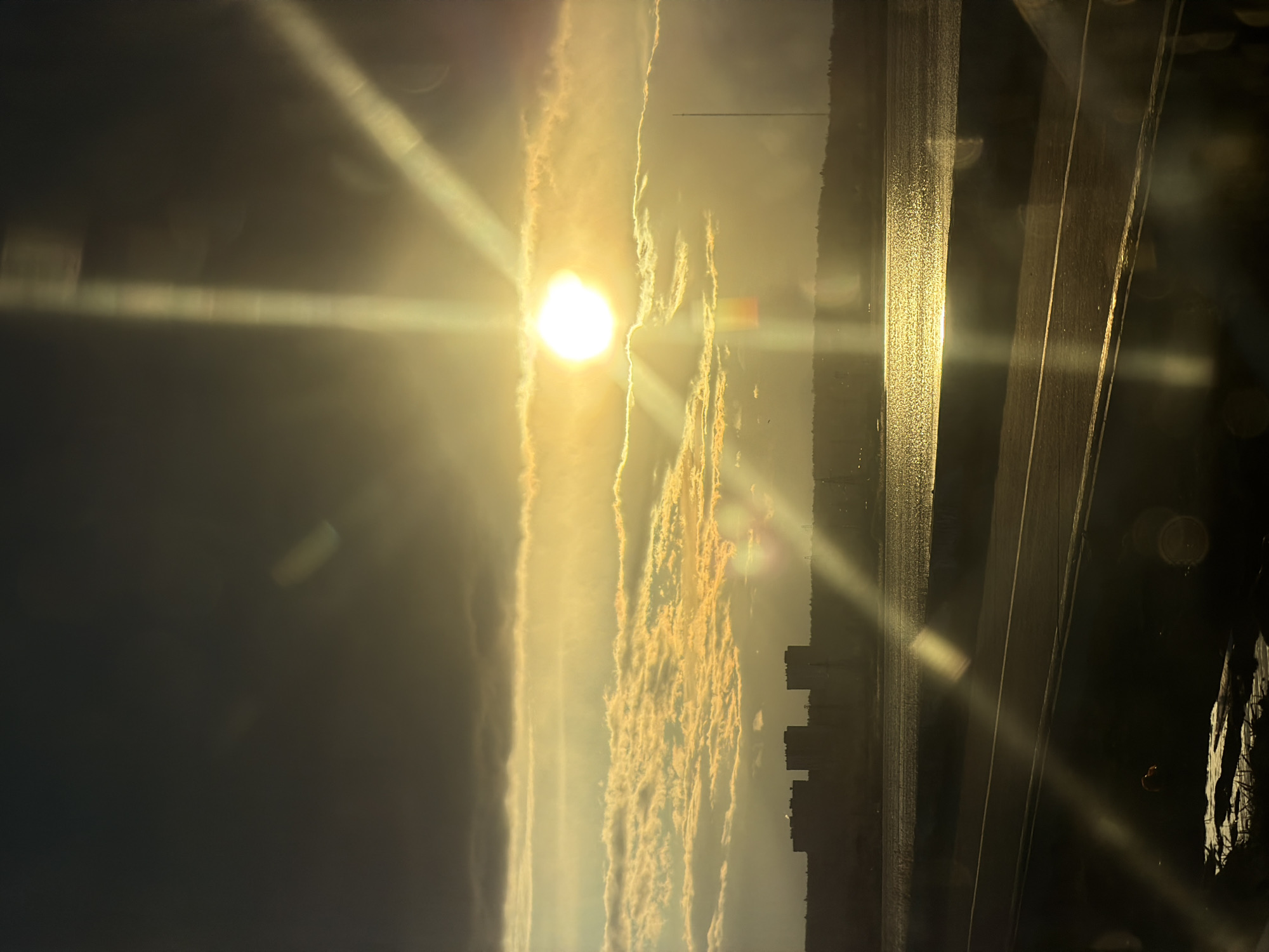 The image depicts a striking sunrise or sunset over a large body of water, likely the Izhevsk Pond, with the distant silhouette of a city skyline. The sun, a brilliant, intensely bright orb, is positioned prominently in the upper-middle of the frame, emitting strong golden light across the sky and water.
Elongated, streaky clouds, illuminated by the sun's warm glow, stretch horizontally across the sky, creating vivid patterns of gold and orange. The surface of the water reflects the sun's radiance, appearing as a shimmering, reflective band in the mid-ground. Along the far horizon, numerous buildings form a dark, understated silhouette of the cityscape.
The foreground is dark and partially obscured, suggesting the perspective is from an elevated position, possibly through a window or from a balcony, with faint structural elements visible. Significant lens flares are apparent, manifesting as horizontal and diagonal streaks of light emanating from the sun, along with circular glare elements. The intense, warm lighting characterizes the scene as either dawn or dusk, evoking a sense of calm and natural beauty. No discernible text is visible within the image.