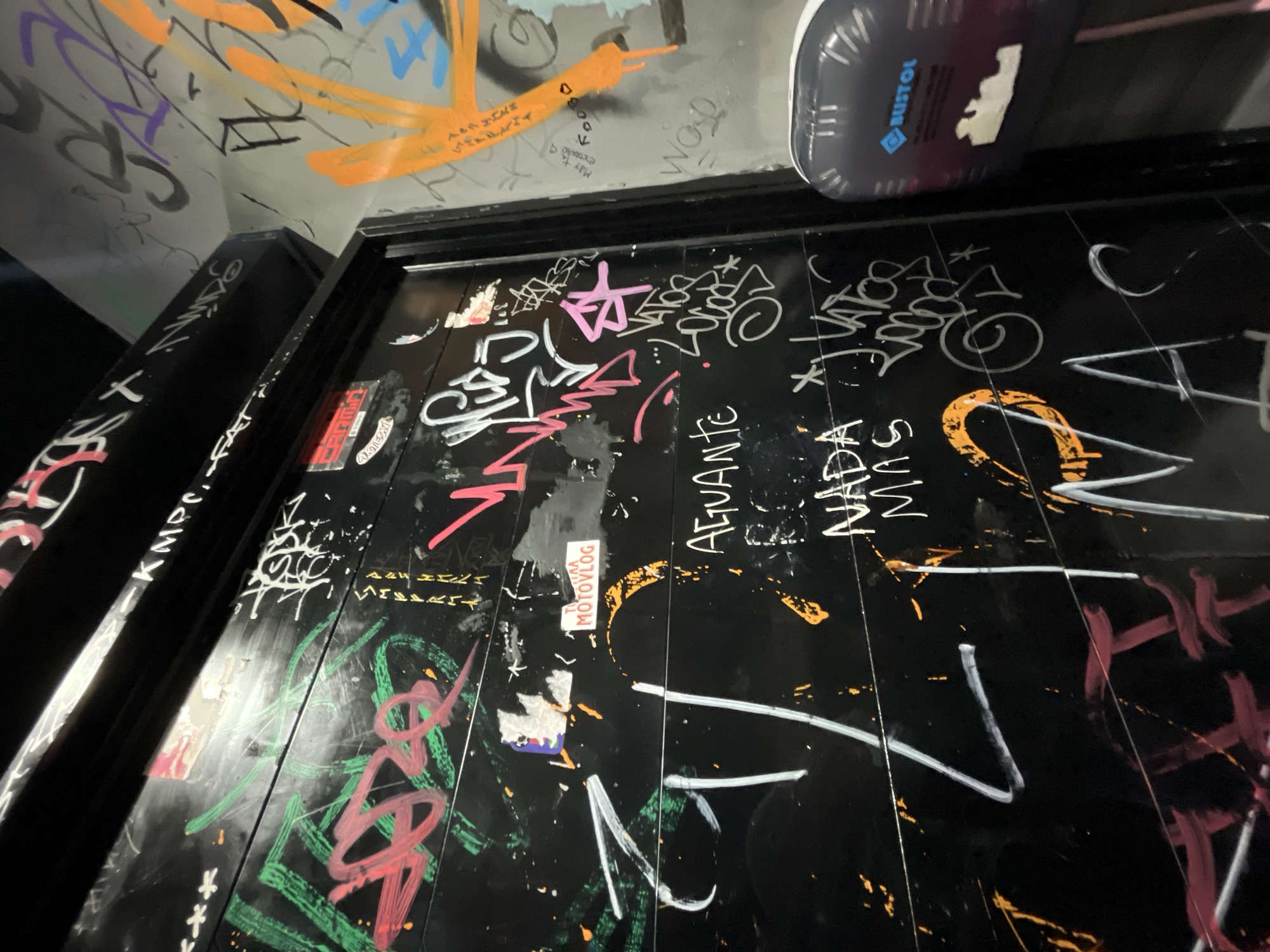This image shows a wall or door covered in graffiti. The writing includes phrases such as "AGUANTE," "NADA MAS," "WOD," and "Lomas de Zamora." There's also a sticker that reads "MOTOVLOG," and the brand "BUSTOL" is visible near the top. The wall or door is black, with colorful graffiti in orange, white, green, pink, and purple. The photograph is taken from a low angle, looking up at the surface. The location, as indicated by the graffiti, is Lomas de Zamora, Argentina.