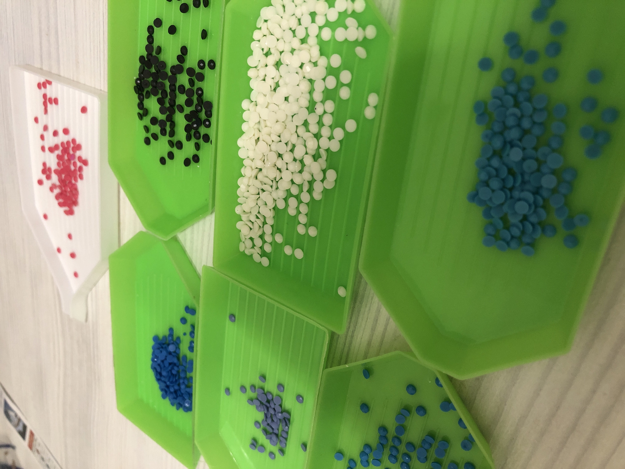 The image shows several small, angled trays containing colorful, round beads. The trays are arranged on a light-colored, textured surface, possibly a table. From left to right, the colors of the beads are: red (in a white tray), black, white, a brighter blue, a darker blue, a light purple and a light blue color again. The trays are mostly green, with the exception of the red beads tray. The beads seem to be sorted by color for some artistic project. No people or any indication of a location in the city of Noviy, Russia are visible.