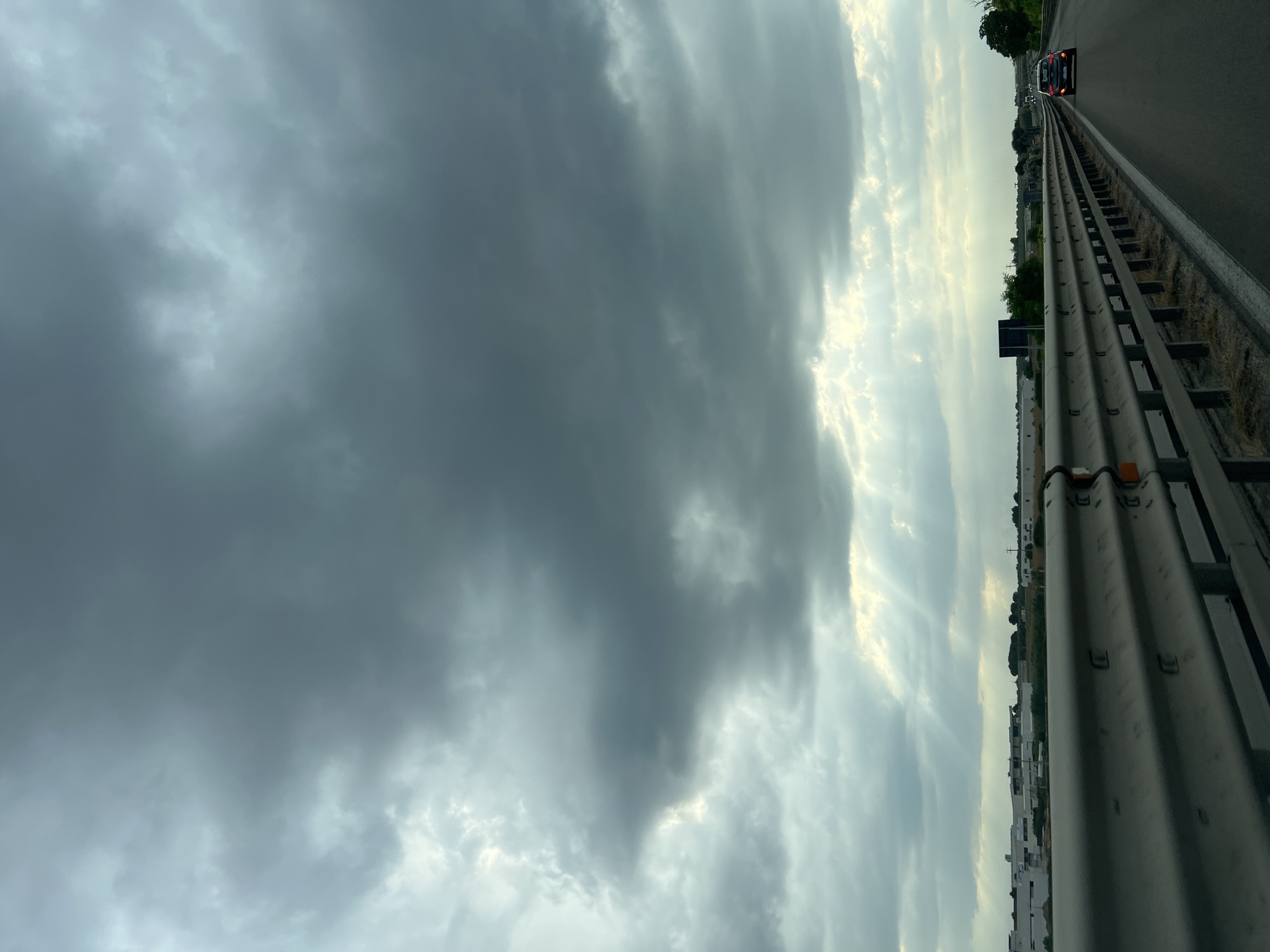 The image captures a view from a vehicle traveling on a highway near Trani, Italy, under a dramatic sky filled with dark, heavy clouds. Sunlight breaks through in scattered rays, creating a dramatic effect against the somber atmosphere. The highway is bordered by a metal guardrail, with dry grass and weeds growing alongside it. In the distance, a flat landscape is visible, featuring industrial buildings with white facades, a few trees, and utility poles. A dark-colored car is the only vehicle clearly visible on the road ahead, traveling in the same direction. The overall impression is one of approaching or receding weather, with a sense of movement along the highway.