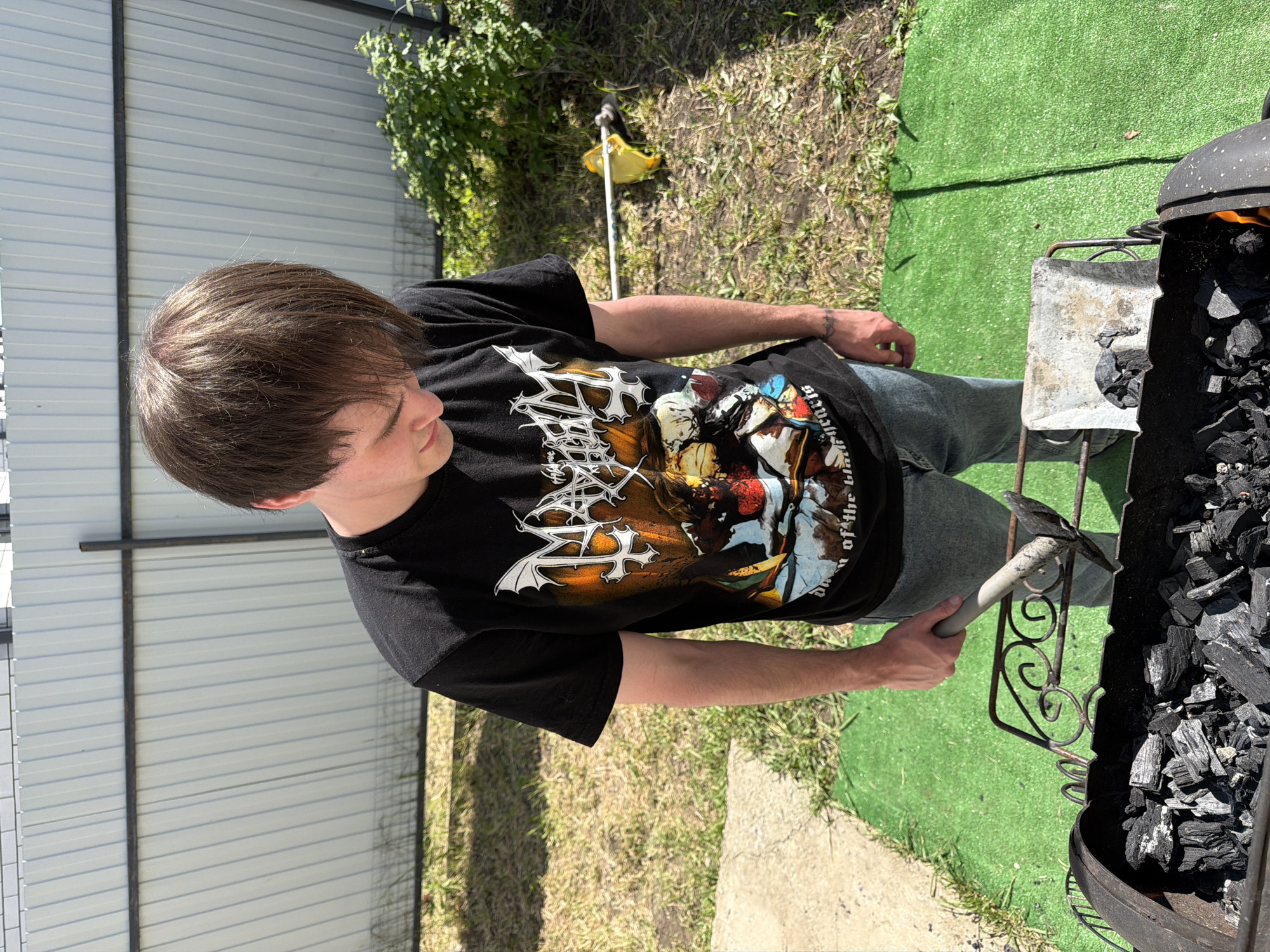 A man stands outdoors, tending to a charcoal grill. The man is wearing a black t-shirt with a graphic design, jeans, and has short brown hair. He holds a metal tool in his right hand, using it to manipulate the charcoal within the grill. The grill is filled with black charcoal, some of which appear to be glowing orange, indicating they are lit. The setting appears to be a backyard or outdoor area, with a section of metal fence in the background. There is also some greenery visible, including grass, weeds and bushes. The location is Rossiyskiy, Russia.