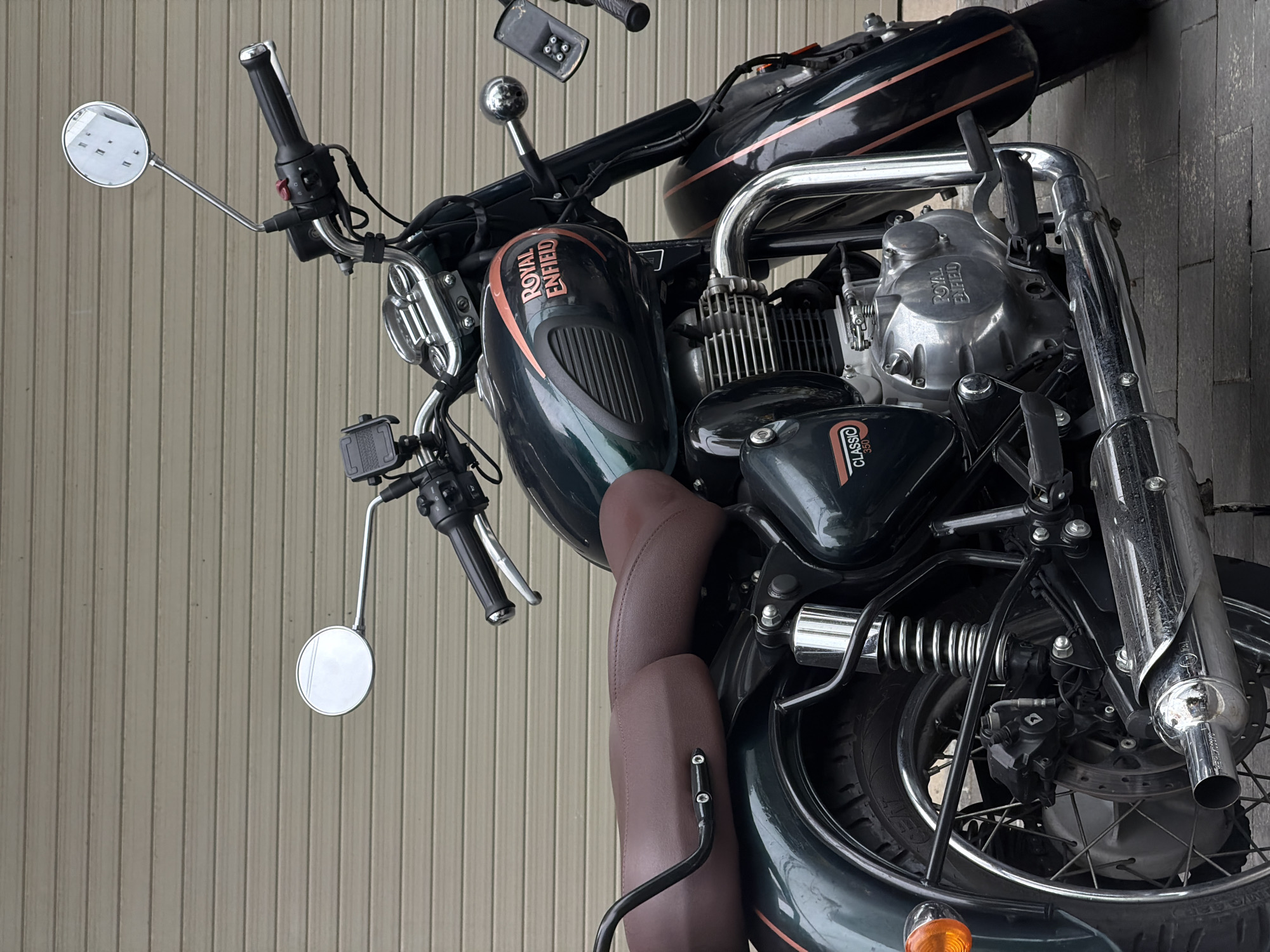 A close-up shot shows a Royal Enfield Classic 350 motorcycle parked against a beige-colored wall with vertical lines. The motorcycle is primarily dark green with copper-colored accents. Visible parts include the fuel tank labeled "ROYAL ENFIELD," the engine block labeled "ROYAL ENFIELD," the seat, handlebar, mirrors, front tire, and chrome exhaust pipe. The license plate or any location-identifying information is not visible. There is no direct evidence to confirm the location within Đà Nẵng, Vietnam, although the style of the wall suggests it could be from the region. No people are visible in the image.