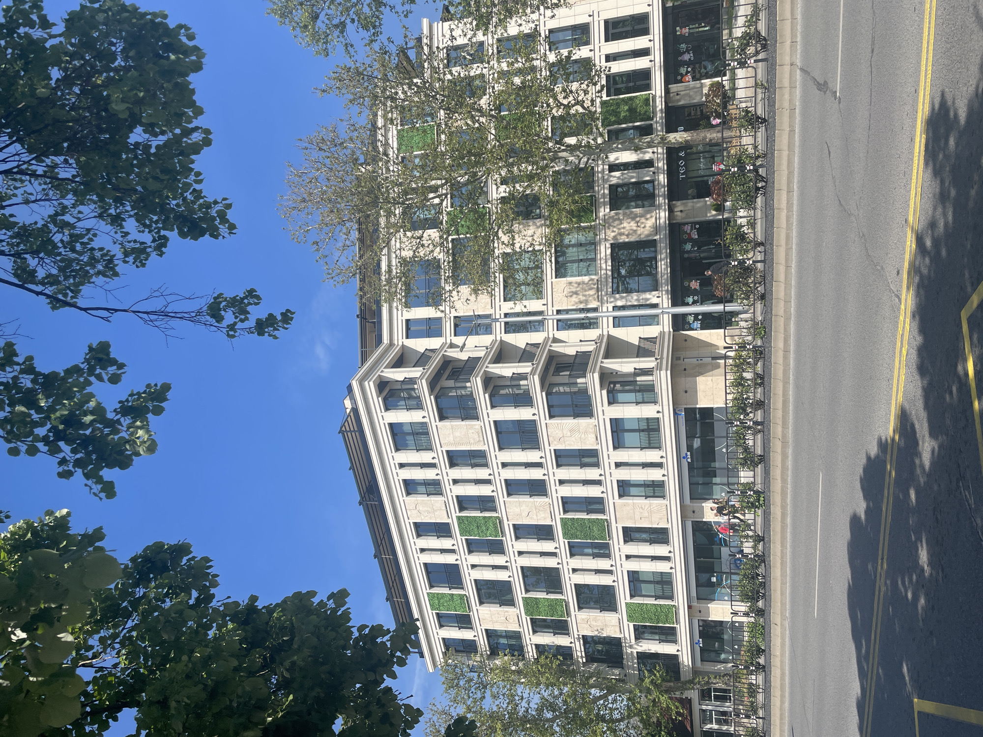 The image, correctly oriented, displays a vibrant urban street scene on a clear, sunny day. The dominant feature is a modern, multi-story building with a light-colored facade, featuring numerous large, rectangular windows. Several sections of the building's exterior incorporate distinctive green panels, appearing as vertical gardens or decorative elements.
The ground floor of the building houses commercial establishments, with visible storefronts. Text on these storefronts includes "TEO &" and "TANGO" on one, and "ДЗМ" on another. Decorative items, possibly floral, are placed outside some shops.
In front of the building, an asphalt street with double yellow lines runs parallel to the structure. A sidewalk separates the building from the road, lined with dark metal railings and planters containing small shrubs or bushes. Several bicycles are parked along these railings. A few individuals are visible on the sidewalk, walking or standing near the shops, suggesting light pedestrian activity.
The scene is bathed in bright sunlight, casting shadows on the street, indicating it is daytime. Large, leafy green trees frame the upper and lower portions of the image against a vivid blue sky, suggesting pleasant weather. The overall impression is of a bustling yet well-maintained street in a city, identified as Plovdiv, Bulgaria.