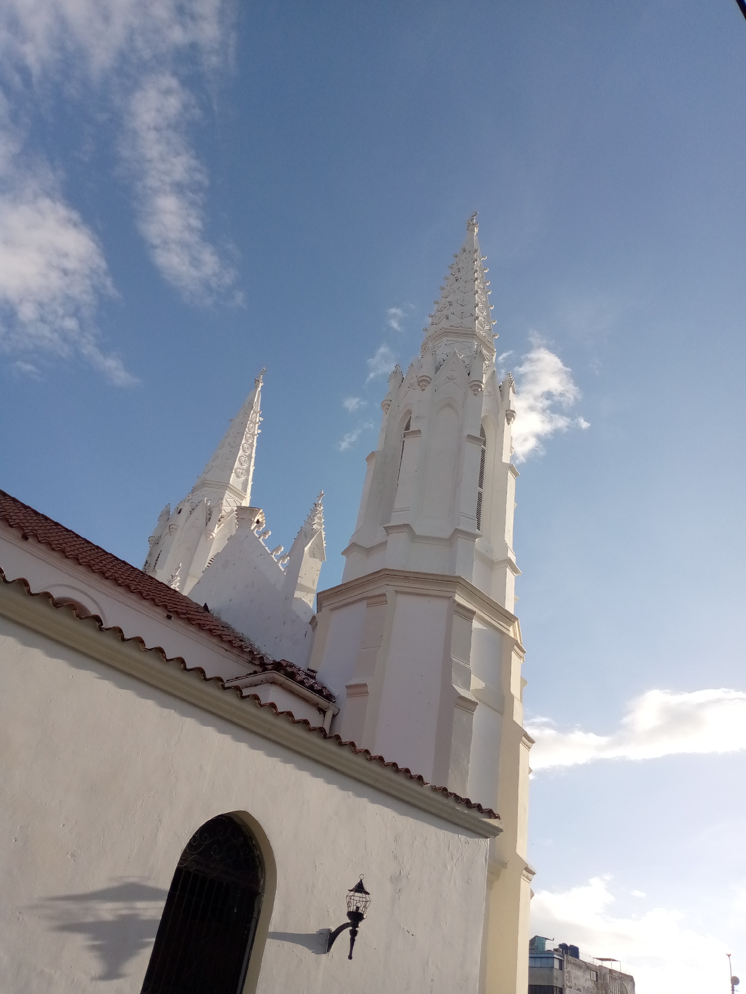 The image captures a low-angle view of the Iglesia de San Juan Bautista in Juan Griego, Venezuela. The church's white facade and intricate spires dominate the frame, set against a bright blue sky dotted with wispy clouds. The red-tiled roofline is visible on the left side of the image, contributing to the colonial architectural style. A dark, arched window with ornate metalwork is present on the lower left wall, alongside a decorative outdoor lamp. In the distance, beyond the church, a modern building with dark-colored water tanks on its roof is partially visible. The scene is devoid of people, emphasizing the architectural grandeur of the church.