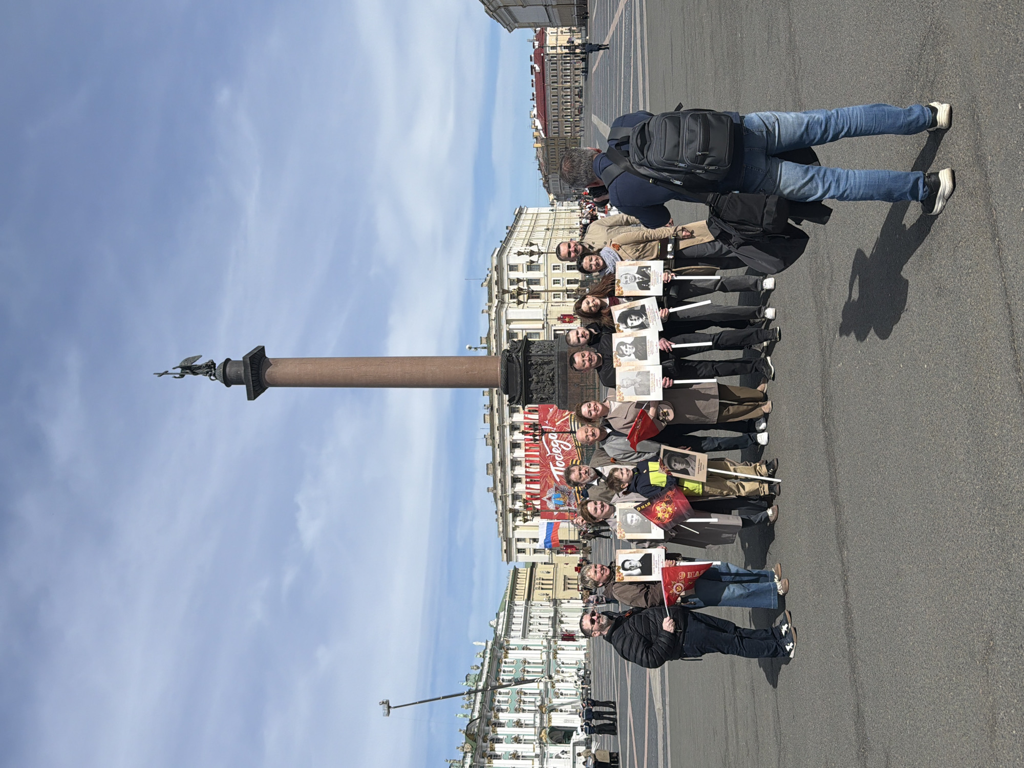 A group of about twelve people stand on a paved area next to the Alexander Column in Saint Petersburg, Russia. They hold photographs of individuals and Soviet red flags. One person is taking a picture of the group. The background includes the Winter Palace and a clear blue sky.