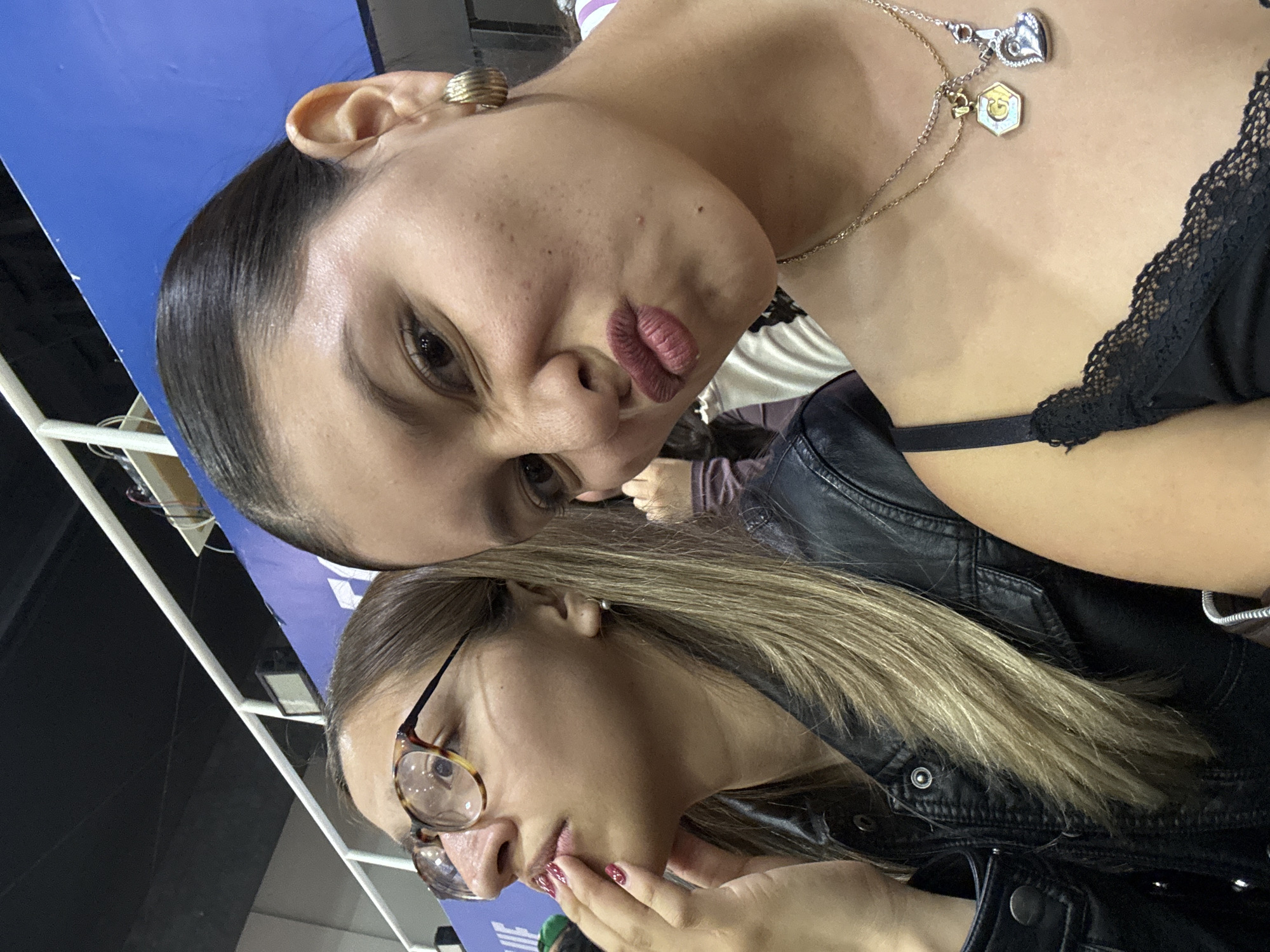 Two women are posing for a selfie indoors. The woman closer to the camera wears tortoiseshell glasses and a black leather jacket. She has light brown hair and her hand is near her mouth. The woman behind her is making a kissing face and wears a black top with lace trim and layered necklaces. She has dark hair pulled back and wears a ring on her ear. The background is partially visible, showing white beams and a blue surface. The location is identified as Heroica Puebla de Zaragoza, Mexico.