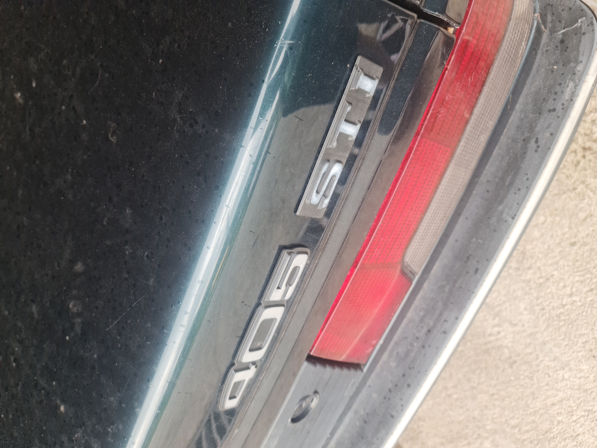 The image captures a close-up, angled view of the rear right section of a dark green car, likely a Peugeot 405. The car's paintwork appears dusty and shows minor scratches, suggesting it has been used or parked outdoors. A prominent rear taillight assembly is visible, featuring a large red section for the brake/running light and a smaller, clear or white section, likely for the reverse light, on its inner edge.
Two car badges are affixed to the car body, to the left of the taillight. The upper badge clearly displays the text "STi" in raised, silver-colored lettering. Below it, a larger badge shows "405", also in raised silver characters, confirming the car's model as a Peugeot 405. Both badges appear slightly worn or discolored around their edges.
The shot is taken during daytime, indicated by the bright, even lighting. No people or active events are depicted. A textured, light-colored surface, possibly concrete or pavement, is partially visible along the bottom right edge of the frame. The overall impression is a static detail shot of a car in an outdoor setting in Buzovgrad, Bulgaria, highlighting its model and trim level.