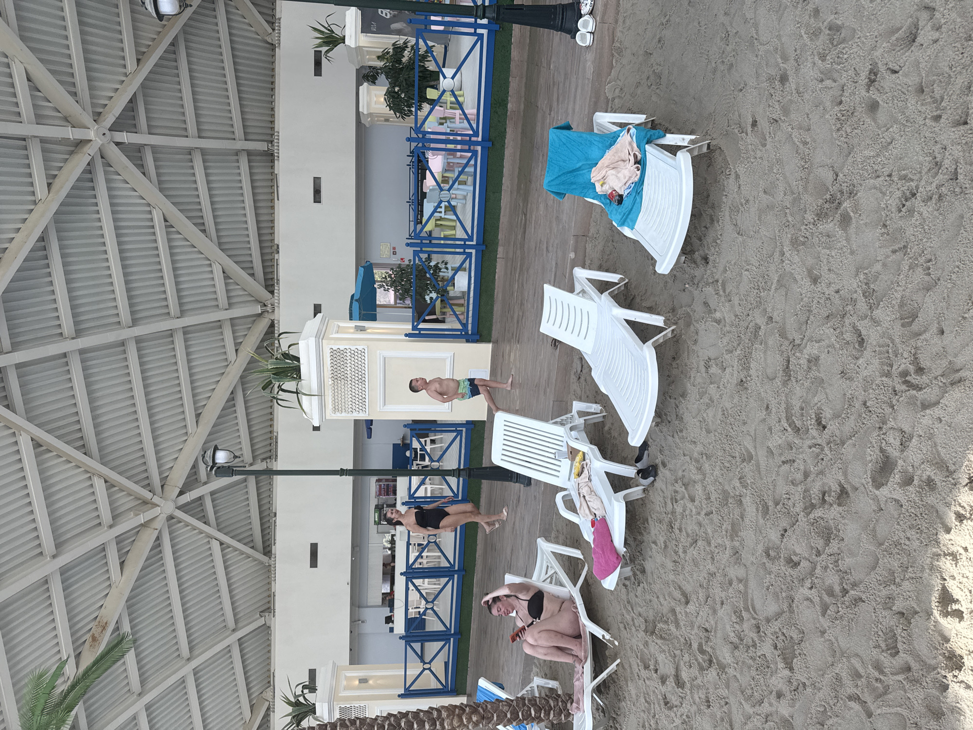 The image depicts a beach resort scene in Estosadok, Russia. In the foreground, several empty white lounge chairs are arranged on a sandy beach. One chair in the center foreground has a blue towel and some clothing draped over it. To the left, a woman in a black bikini is sitting on a lounge chair, looking at her phone. Further back, a woman in a black swimsuit walks away from the camera, and a young boy in swim trunks runs towards the right. Behind them, a blue railing encloses an area with tables and chairs, possibly a cafe or dining area. The resort has a large, covered structure with a metallic roof supported by beams, and palm trees are visible on the left and right sides of the image. The overall atmosphere suggests a leisure setting, likely at a hotel or resort.