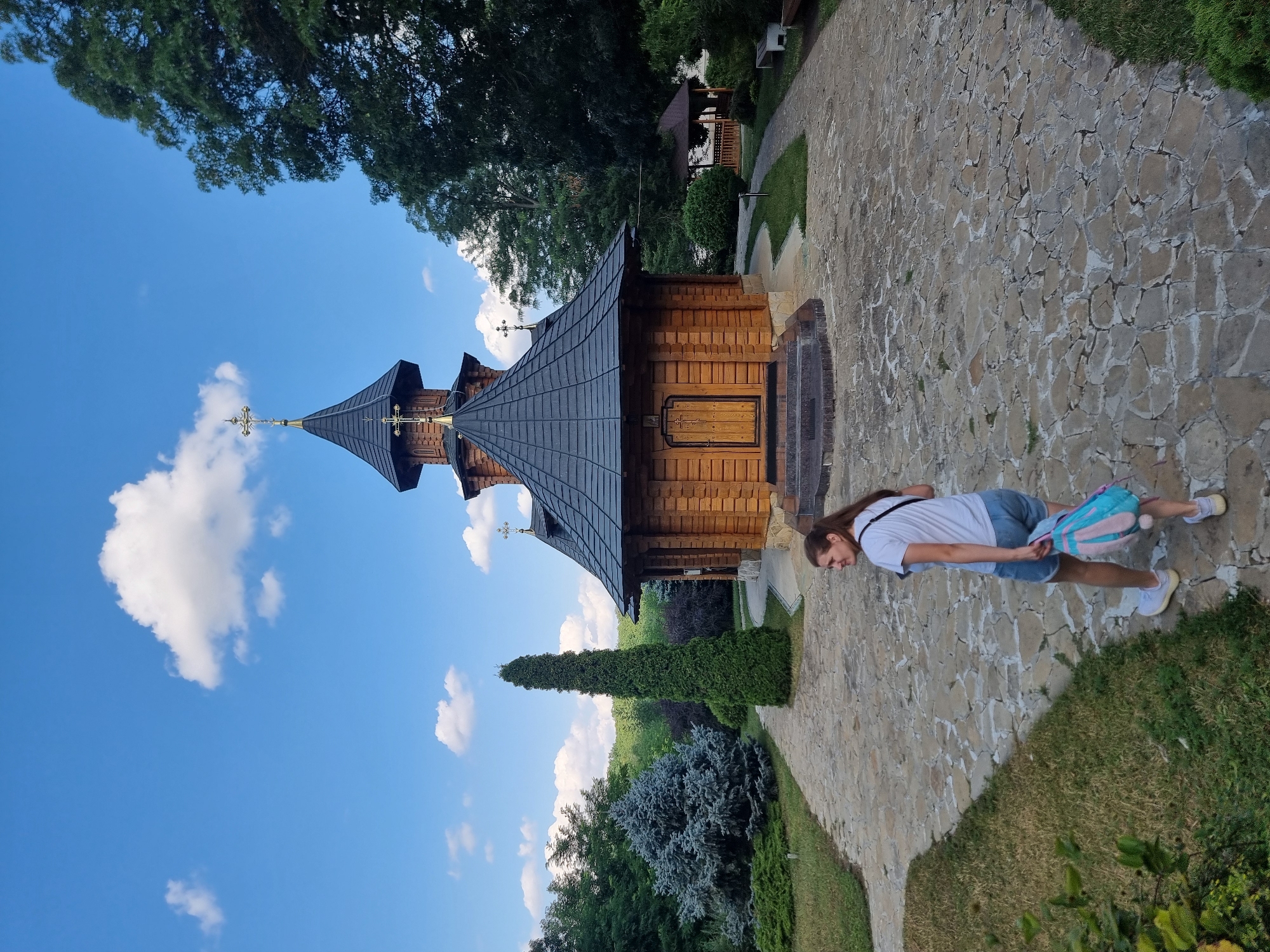 A young woman with long brown hair is standing on a stone pathway next to a wooden church. She is wearing a white t-shirt, blue denim shorts, and white sneakers. She is carrying a backpack with blue, pink, and white patterns. The church is a traditional Orthodox wooden structure with a dark grey, multi-tiered roof topped with golden crosses. The church is situated in a lush green setting with trees and bushes. The sky is bright blue with scattered white clouds, indicating it is daytime. The weather appears to be pleasant and sunny. The scene is likely outdoors at a religious site. The image was taken in Hagimus, Moldova. There is no visible text in the image.
