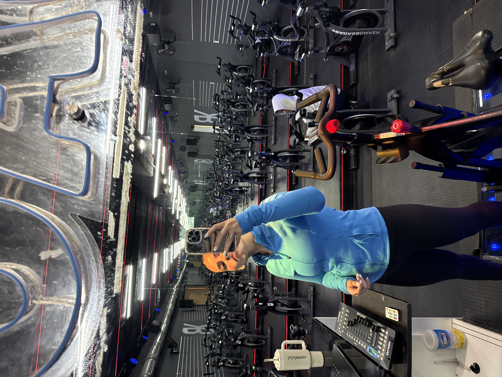 A person with blonde hair is taking a selfie in a spin class studio. The individual is wearing a light blue jacket and dark leggings. The studio contains rows of stationary bikes. A sound mixing board, a bottle of Clorox wipes, and a white water bottle labeled "momslulul" are on a shelf in the foreground. The text on the wall reads "#BYOBBIKER." The studio is located in the city of Puebla, Mexico.