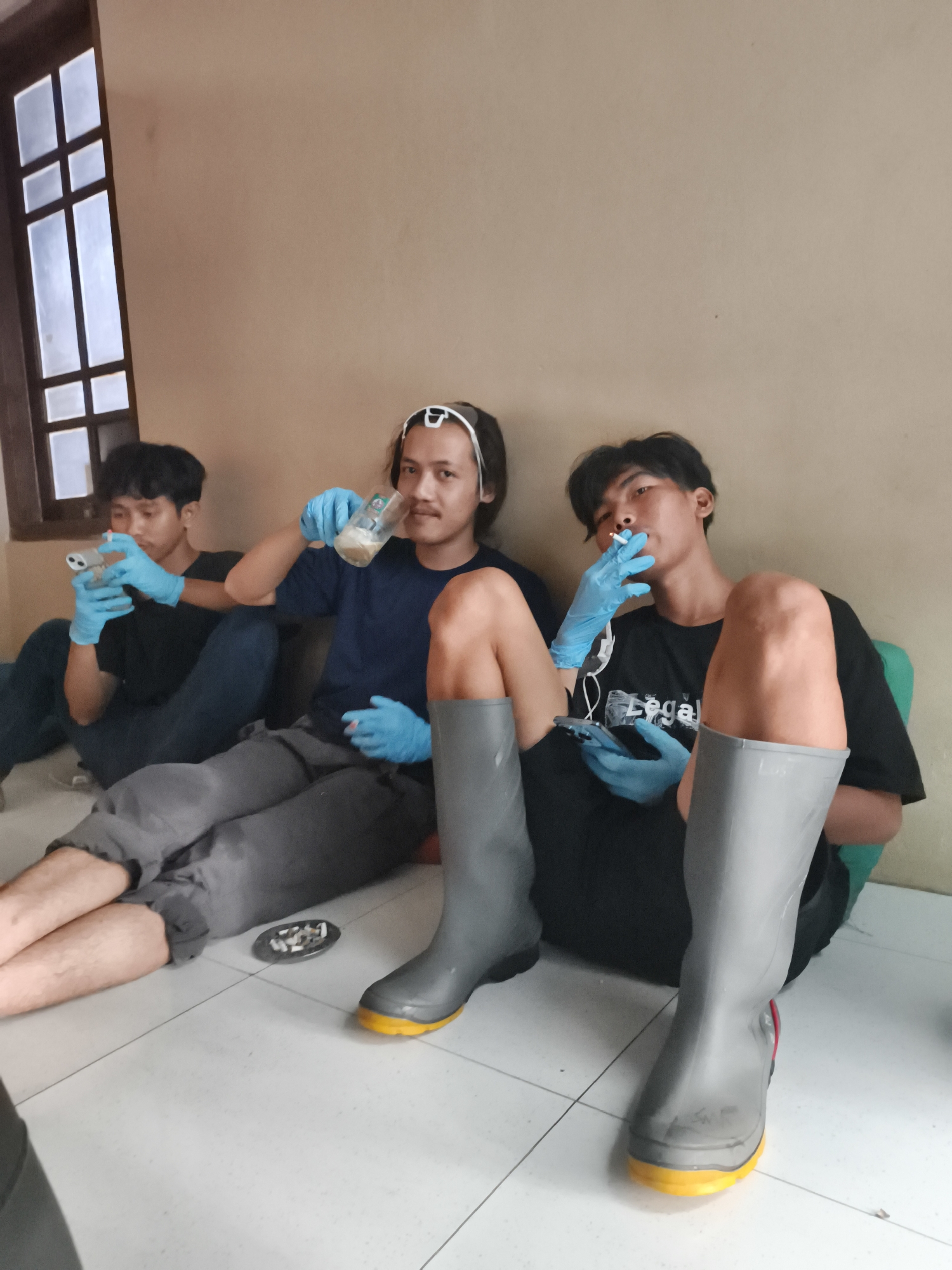 In Pekalongan, Indonesia, three young men sit on a tiled floor against a tan wall, near a window with wooden mullions. The man on the left, wearing a black t-shirt and blue jeans, is looking at his phone, also wearing blue gloves. The central figure, in a dark blue t-shirt and grey pants, holds a glass of light brown liquid and wears blue gloves. The man on the right, in a black t-shirt, is smoking a cigarette and holding a phone, also wearing blue gloves. All three men are wearing grey rubber boots. An ashtray filled with cigarette butts rests on the floor between the man in grey pants and the man in the black t-shirt.
