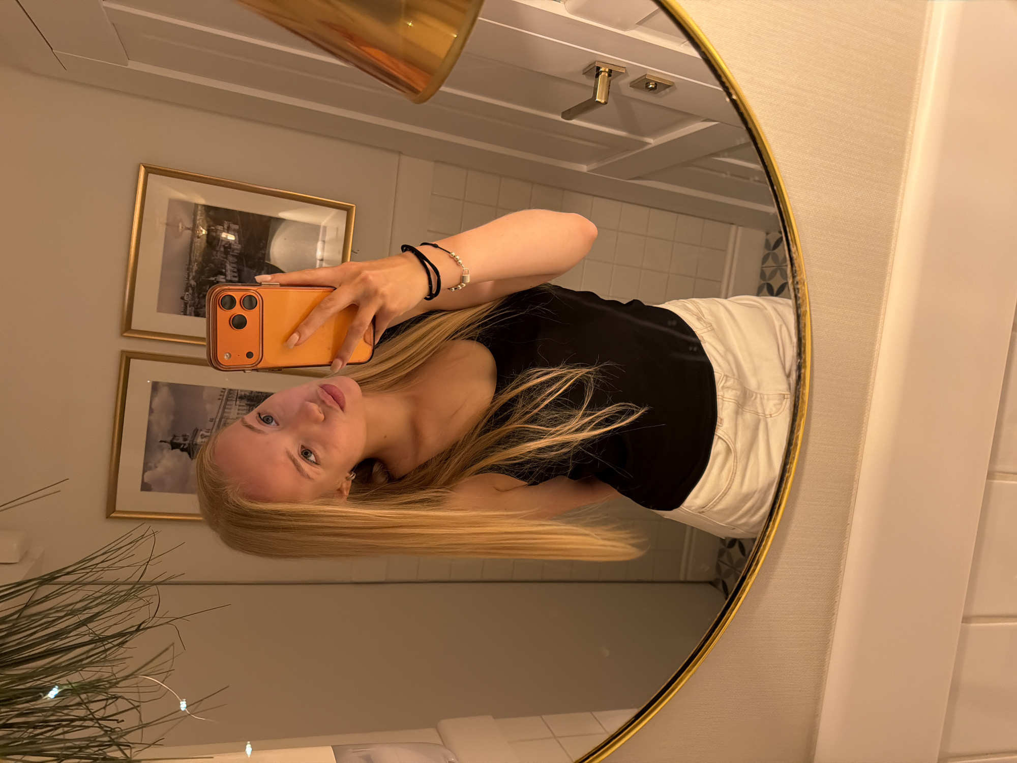 A woman is taking a selfie in a bathroom. The woman has long blonde hair, and she is wearing a black shirt and white pants. She is holding an orange smartphone in her right hand. The bathroom has a round mirror with a gold frame. There are two framed pictures on the wall behind her. A light fixture is visible at the top of the mirror's reflection. A plant is on the left side of the mirror. The specific city and state location are unknown.
