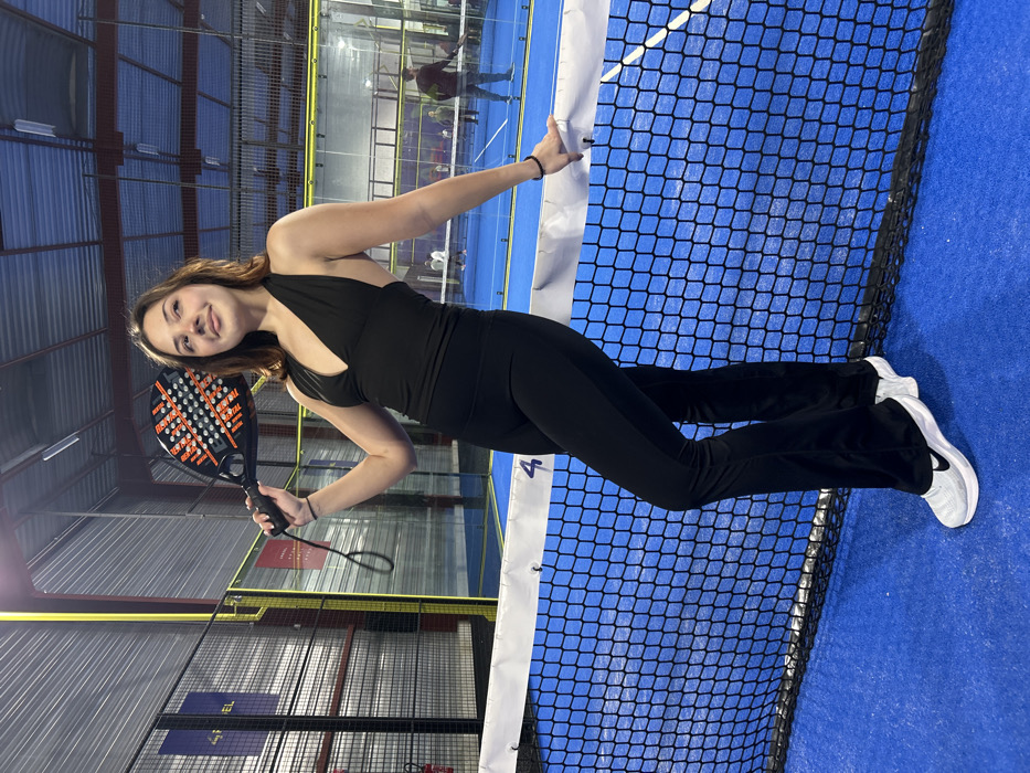 An indoor portrait displays a young woman standing upright on a blue padel tennis court, smiling directly at the viewer. She is positioned next to the net, which spans horizontally across the image. The woman wears a black halter-neck sports top, black flared athletic pants, and white athletic shoes with a black Nike swoosh logo. In her right hand, she holds a black and orange/red padel racket, displaying the brand "SIUX" on its surface. Her left hand rests casually on the white band of the padel net.
The padel court features transparent glass walls, typical of the sport, through which blurred figures of other people are visible in the background, suggesting an active sports complex environment. The ceiling reveals industrial-style lighting fixtures, indicating an indoor setting, likely in the evening or at night. Metal support structures for the court enclosure are also visible. On the white band of the net, a number "4" is partially discernible, possibly indicating the court number. Further in the background, fragmented text "FEL" can be seen on a wall or banner. The scene captures a moment of leisure or post-activity on a padel court in Eckbolsheim, France.