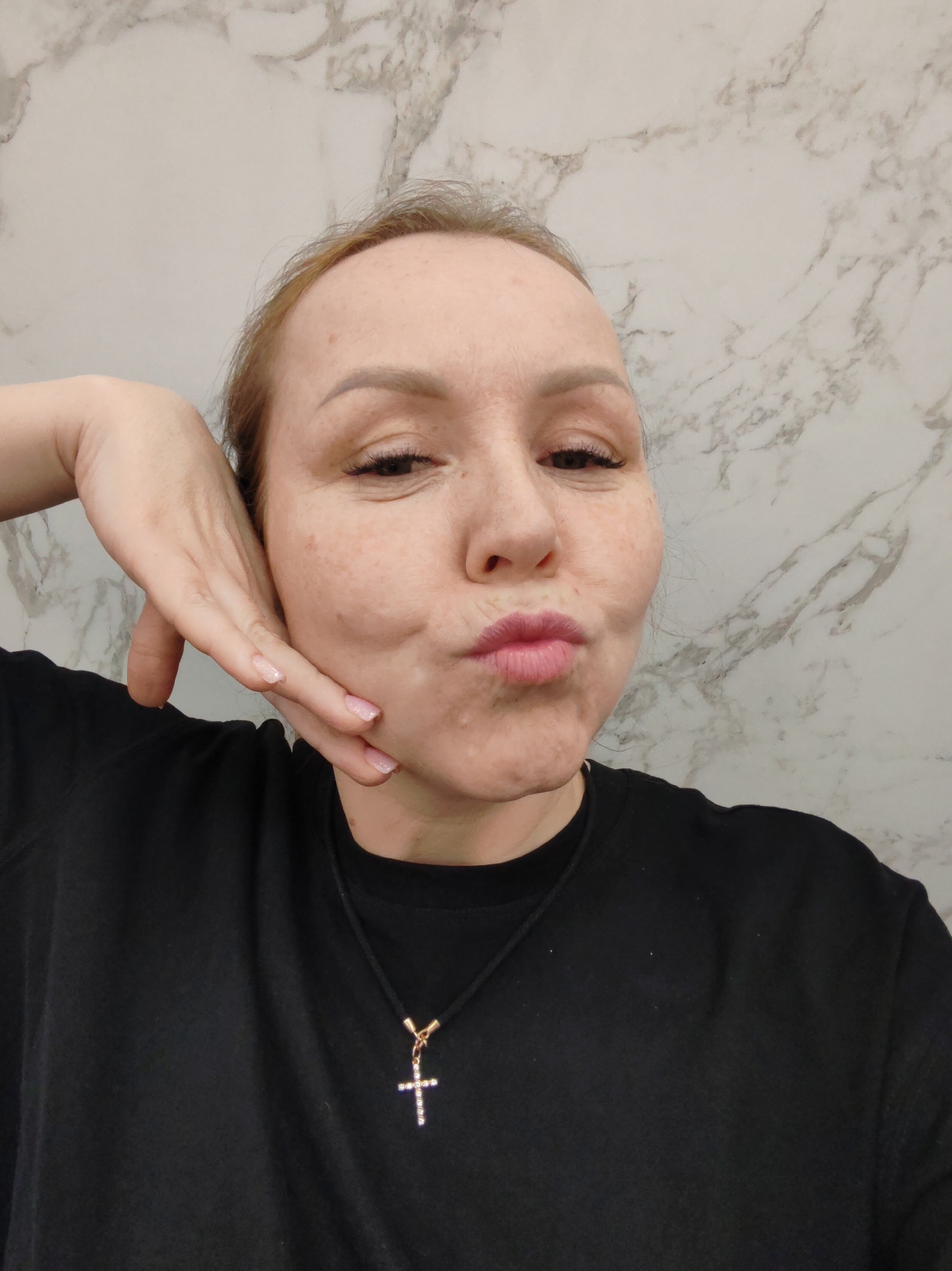 A light-skinned woman with freckles is posing for a selfie. She is wearing a black t-shirt and a necklace with a cross pendant. She is holding her hand up to her face and puckering her lips. The background is a wall with a marble pattern. The image was taken in an indoor location, potentially in the city of Chaikovsky, Russia.