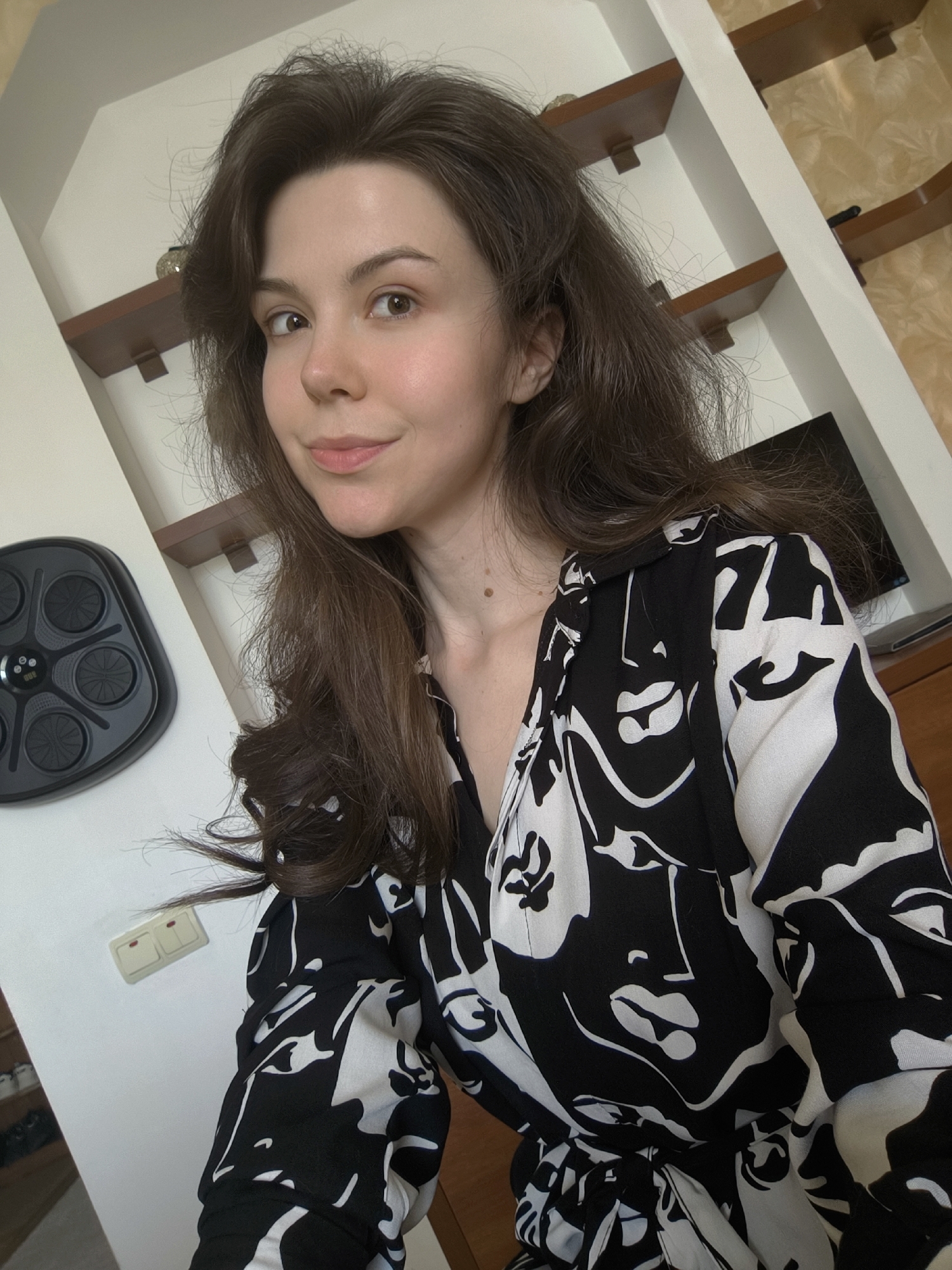 A medium shot shows a young woman with long, dark brown hair taking a selfie indoors. She is wearing a black and white patterned dress. The background includes a white wall with wooden shelves, a television, and a black electronic device on the wall. There is also a light switch on the white wall.