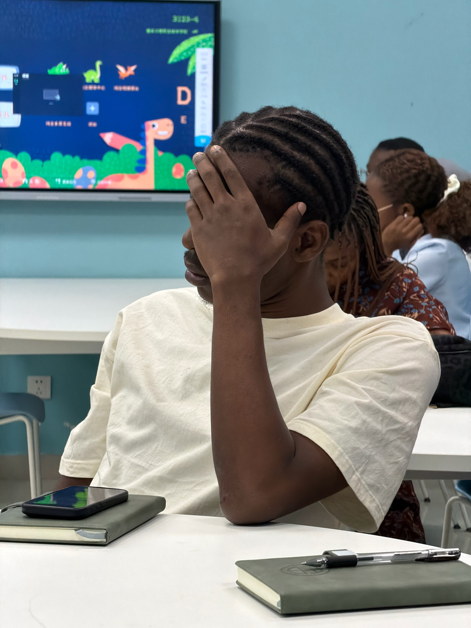 A young Black male with braided hair sits at a white table, covering his eyes with his right hand. He wears a light-colored t-shirt. In front of him, a black smartphone rests on a green notebook. To the right, a pen lies on another green notebook. In the background, a large screen displays colorful animated characters, possibly from an educational program. Several other individuals are visible in the background, seated at tables. The environment appears to be a classroom or lecture hall, characterized by white tables, chairs, and a light blue wall. This scene is likely taking place in the city of Jiangjin, China, given the context provided.