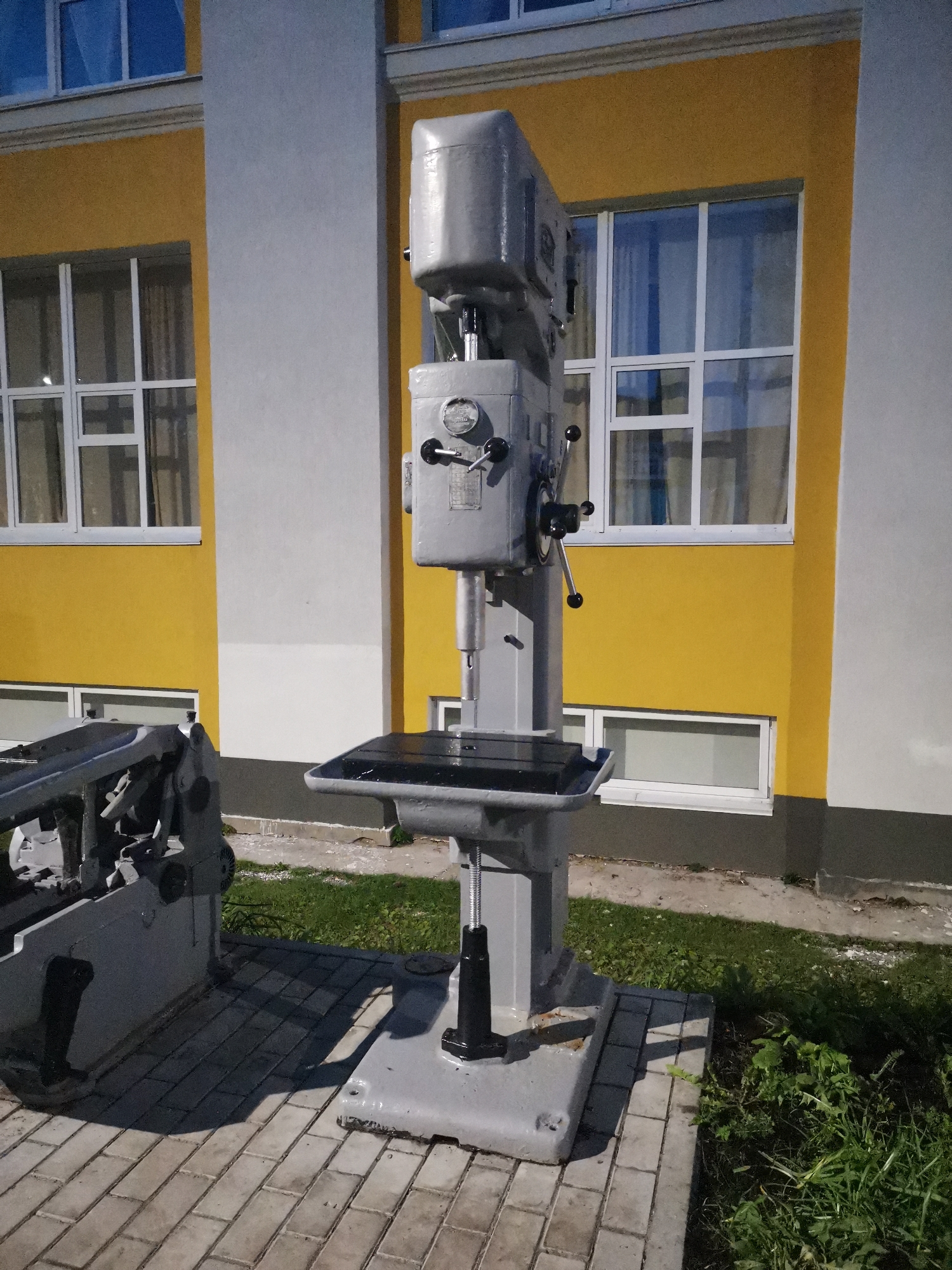 This image shows a large, industrial gray drill press standing on a paved area in front of a building with yellow and white walls and several windows. The building appears to be a school or administrative building. To the left of the drill press is another piece of industrial equipment, also gray. The ground is paved with light-colored rectangular stones, and there is a patch of green grass to the right of the paved area. The lighting suggests it is daytime, possibly late afternoon or early morning due to the shadows. The image is taken from a slightly low angle, looking up at the drill press and the building. The drill press is the central focus of the image. The environment is clean and orderly. It is a still, outdoor scene. This scene is located on the territory of the Kizel Branch of Perm State Technical University in Kizel, Russia.