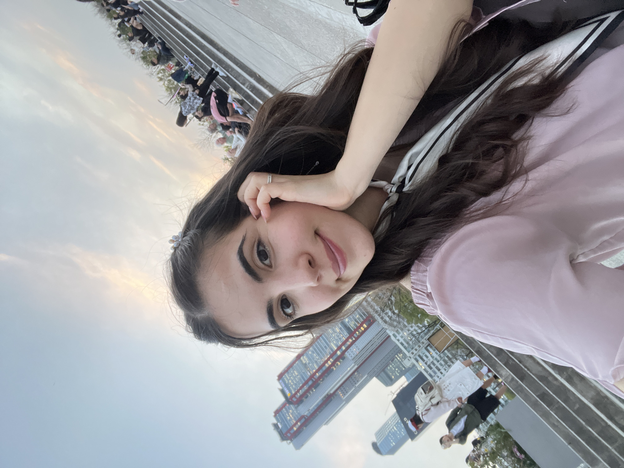 A selfie, taken from a low angle, shows a young woman with long brown hair, fair skin, and dark eyes, wearing a light pink top. Her right hand is near her face. In the background, there are several skyscrapers, and a staircase with a crowd of people. The location is identified as Seoul, South Korea.
