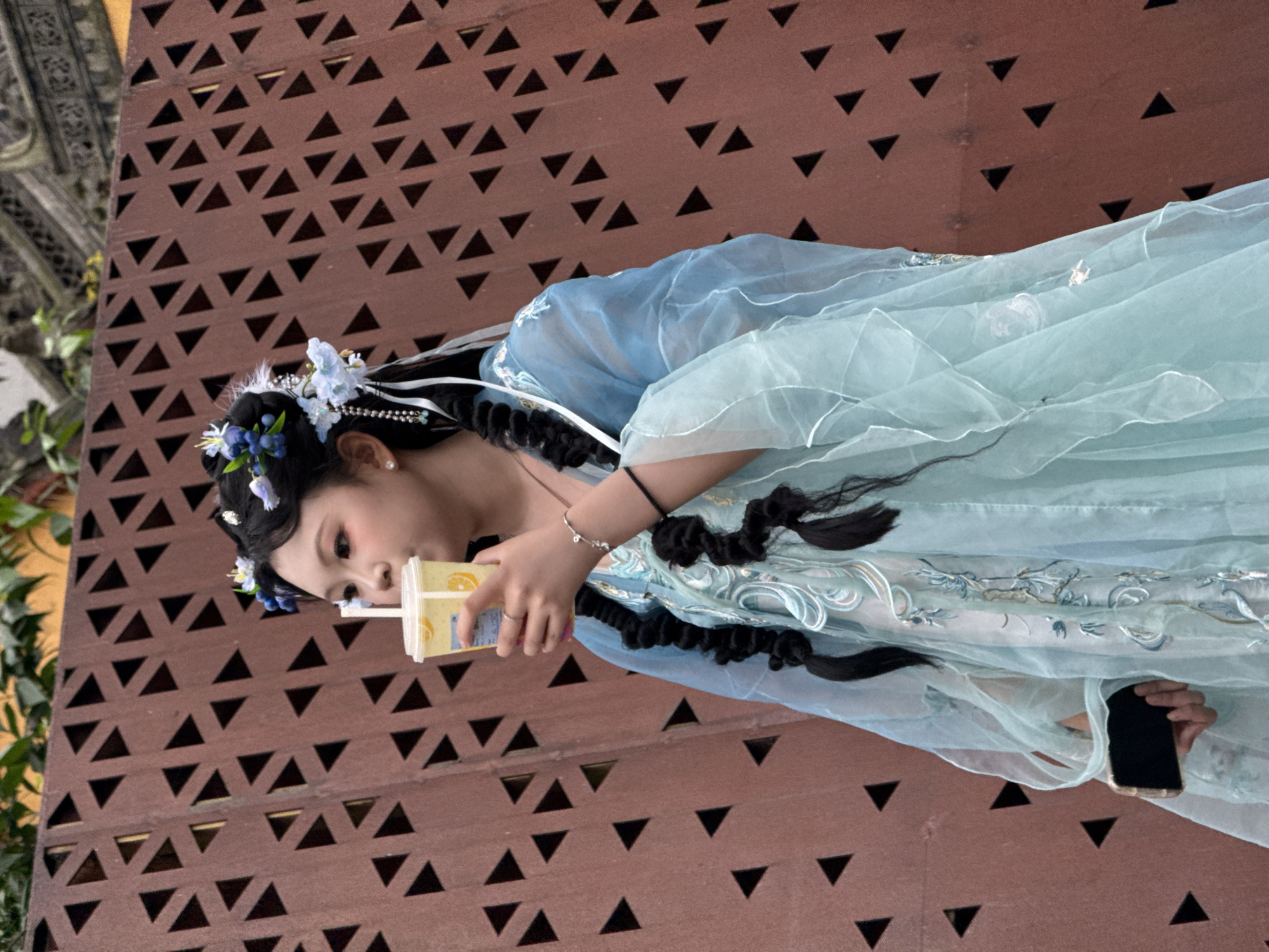 A woman in traditional blue garb stands in front of a rust-colored wall with repeating triangular patterns. She sips from a yellow drink and holds a black smartphone. The location is the Yuzhong District in Chongqing, China.