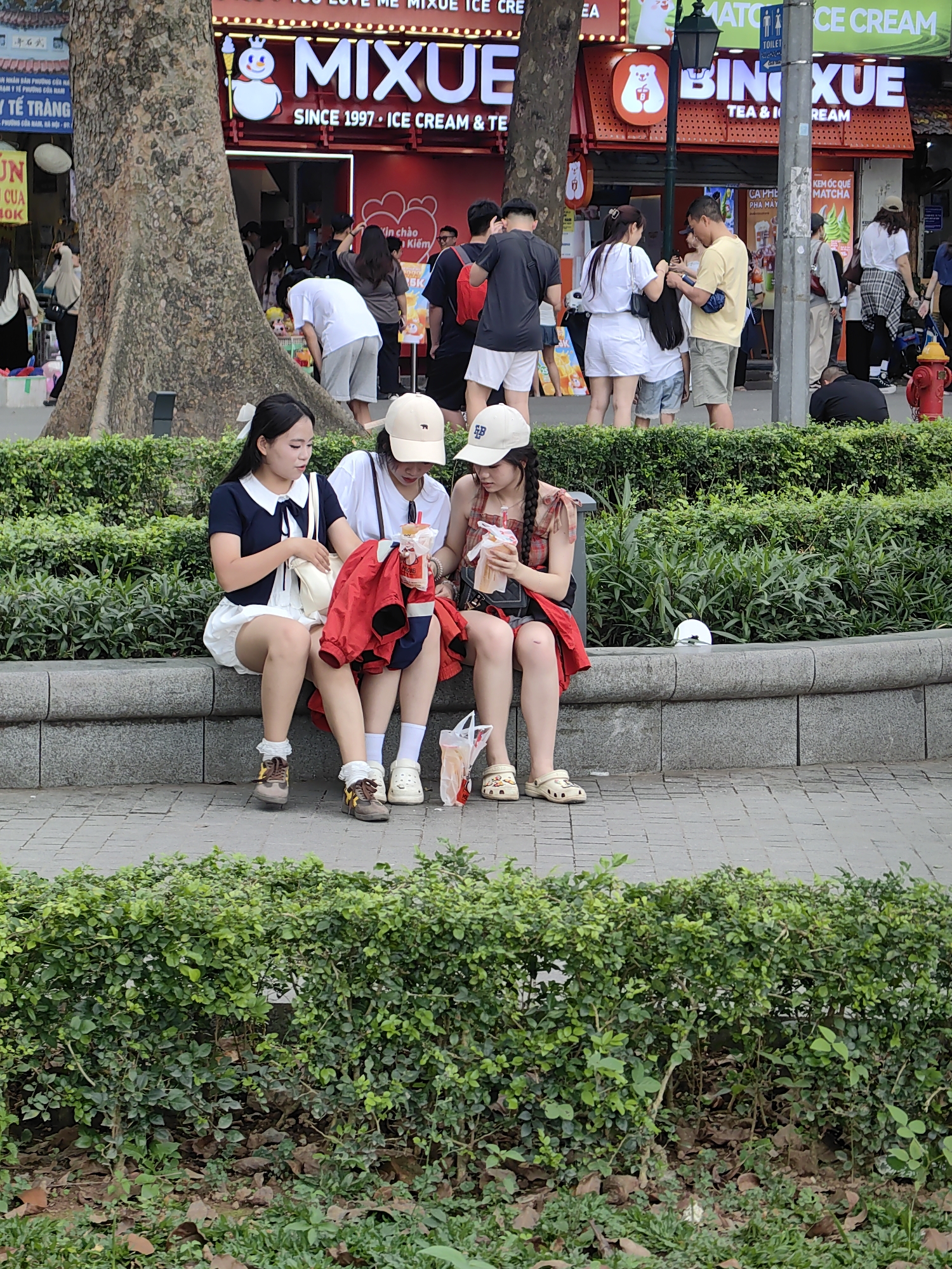 Three young women are sitting on a concrete bench in a public area of Hà Nội, Vietnam. The woman on the left is wearing a dark blue top with a white collar, white shorts, and brown shoes with white socks. The woman in the middle has on a white T-shirt, white shorts, a red jacket, and tan shoes. The woman on the right is wearing a plaid top, dark shorts, a red jacket, and light-colored Crocs. They are near a Bing Xue Tea & Ice Cream shop. People are walking in the background. The foreground features trimmed green bushes.